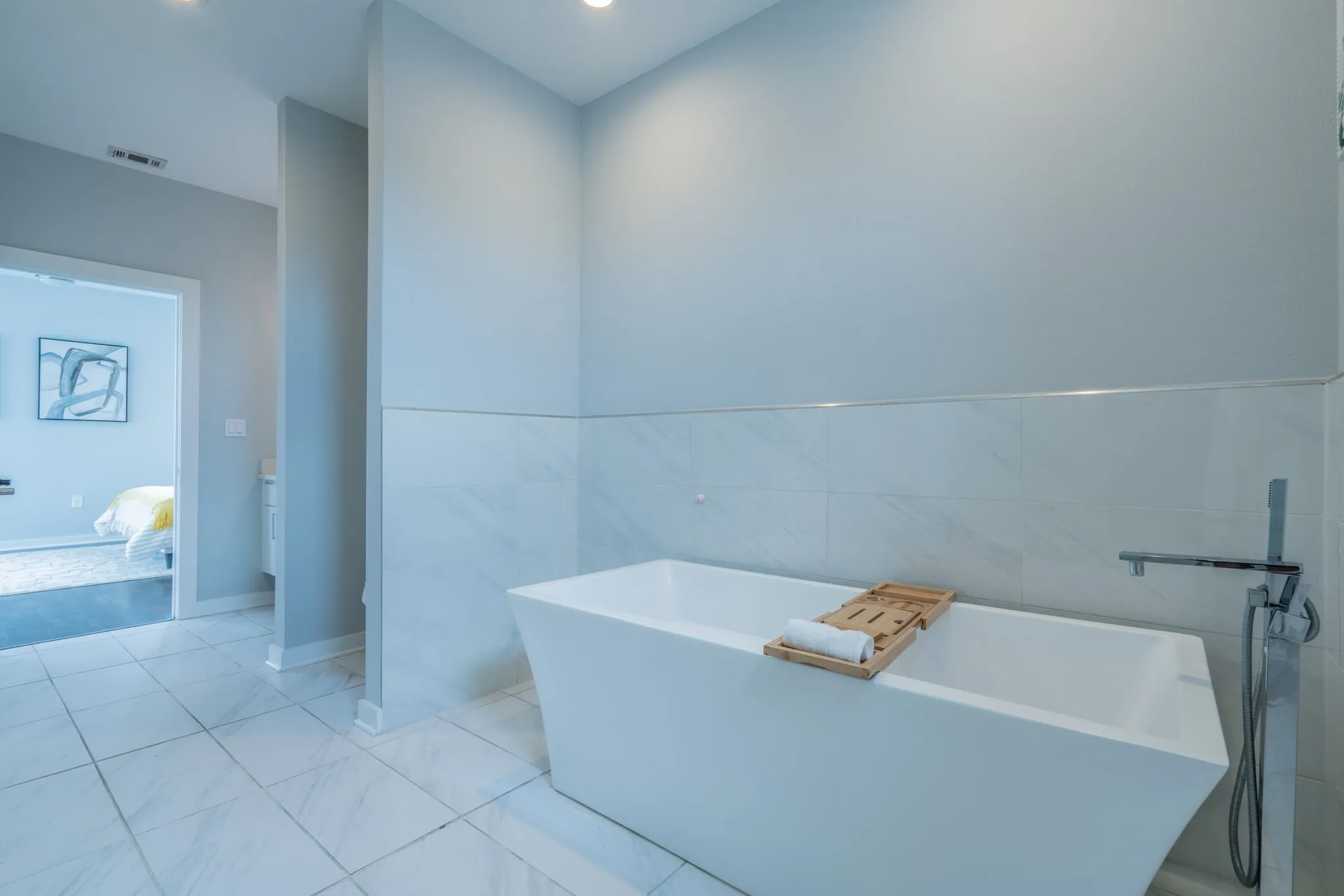 White tiled flooring, freestanding soaking tub, wall-mounted faucet, recessed lighting, and tiled wall accents