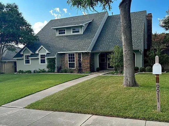 Light green siding, brick facade, dormer windows, and a concrete pathway leading to the entry