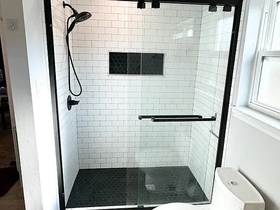Frameless glass shower enclosure with matte black hardware, white subway tile surround, dark hexagonal floor tiling, and a recessed shower niche