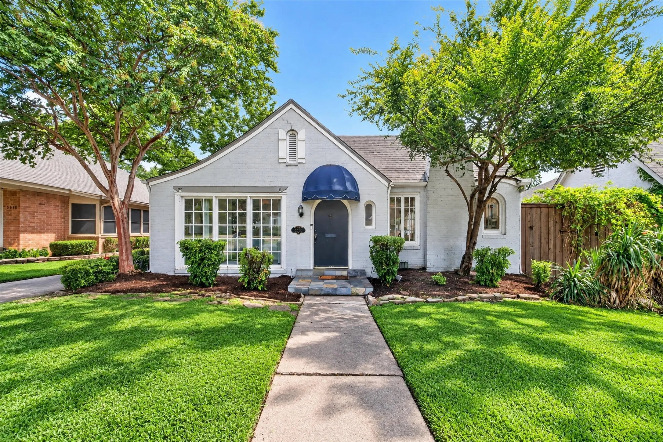 Gabled roofline with stucco exterior, multiple arched windows, prominent front entry with blue awning, and mature trees framing the property