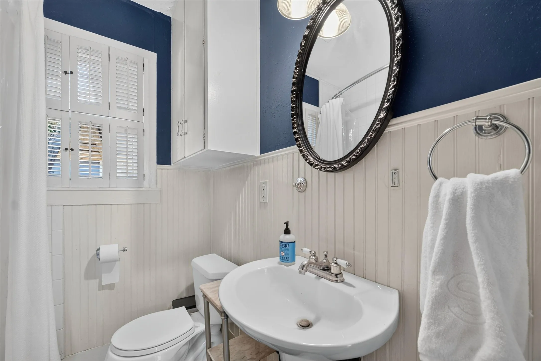 Pedestal sink with chrome fixtures, beadboard wainscoting, wall-mounted cabinetry, window with plantation shutters, and an oval vanity mirror