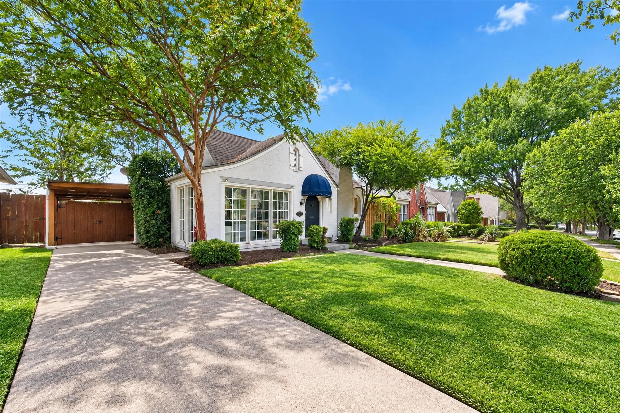 White stucco exterior with a blue awning, expansive front lawn, mature trees, and a concrete driveway leading to a covered carport