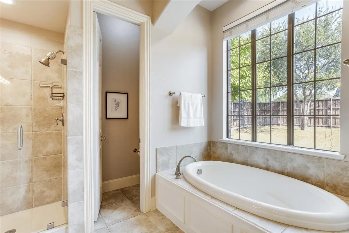 Built-in soaking tub, tiled tub surround, large window with grid pattern, tiled shower enclosure with glass door, and brushed nickel fixtures