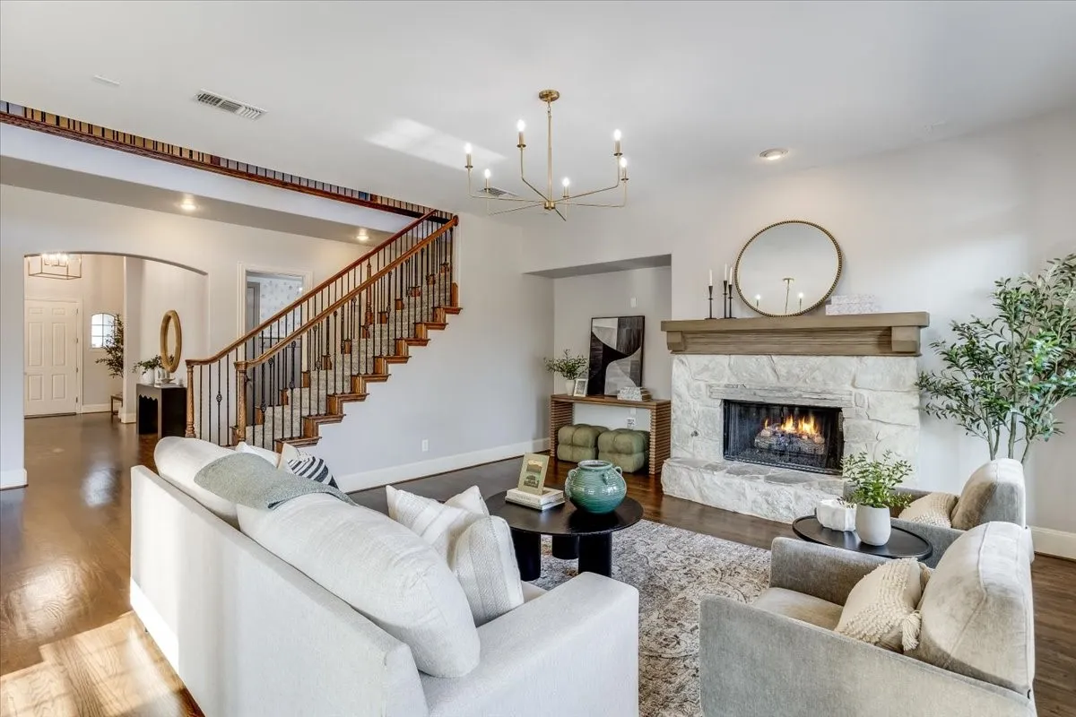 Hardwood flooring throughout, a stone-clad fireplace with a wood mantle, a grand staircase with dark wood railings and wrought iron balusters, arched entryways, and a modern chandelier
