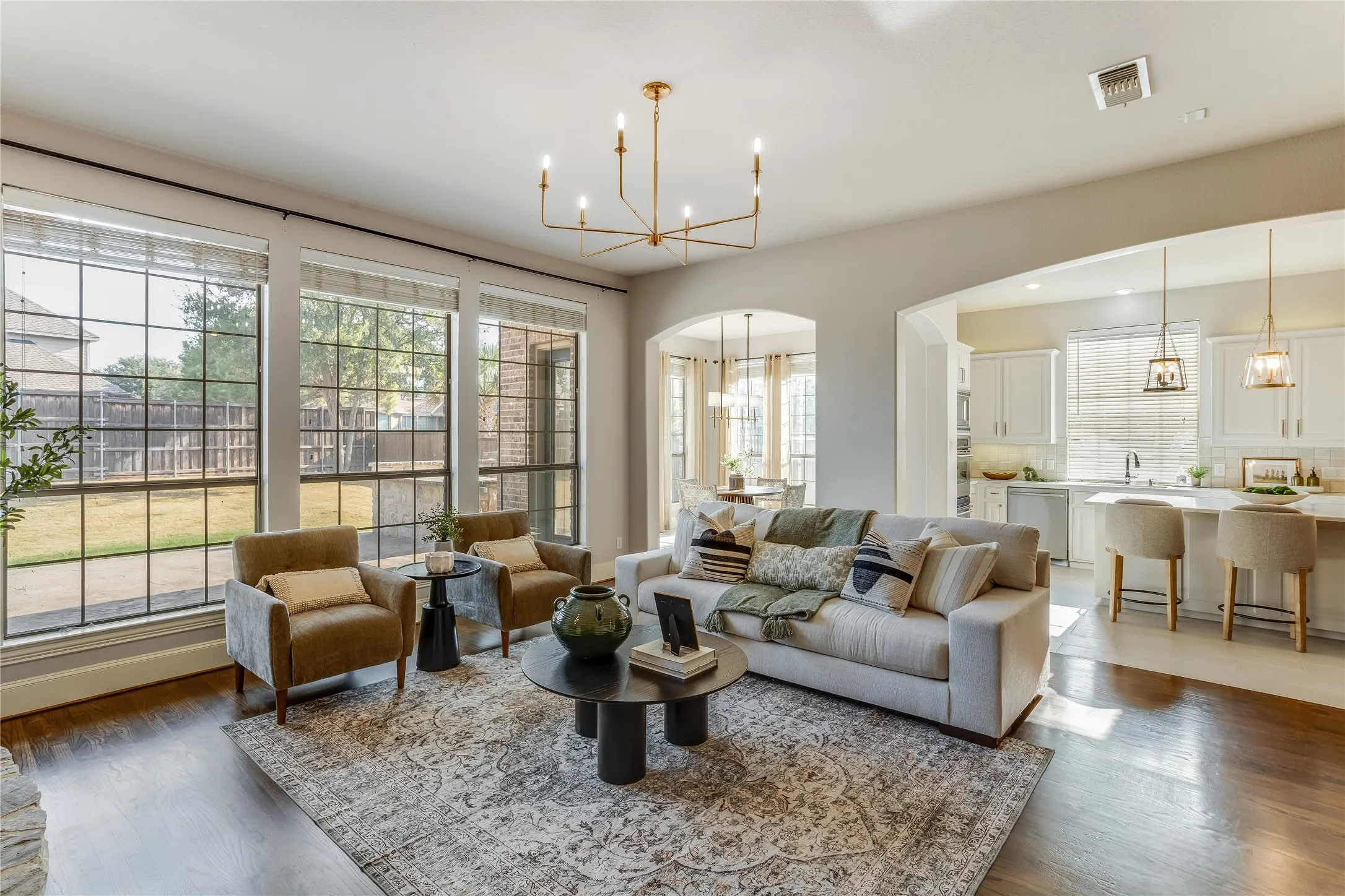 Spacious living area featuring large picture windows, wood-finish flooring, a contemporary chandelier, and an arched doorway to a white cabinet kitchen with a breakfast bar