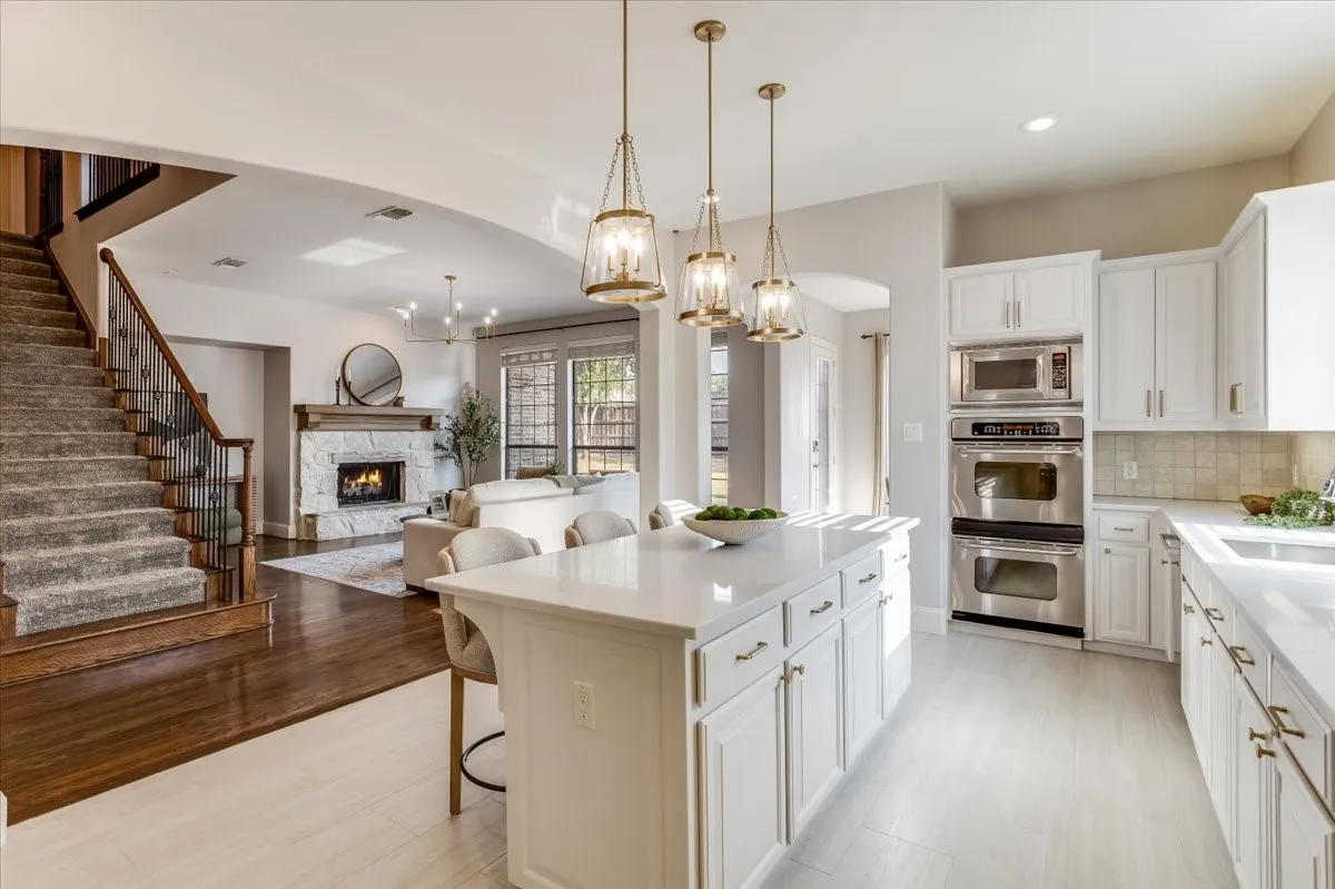 White cabinetry with brass hardware, a central island with countertop seating, stainless steel double ovens, a built-in microwave, and a stone fireplace