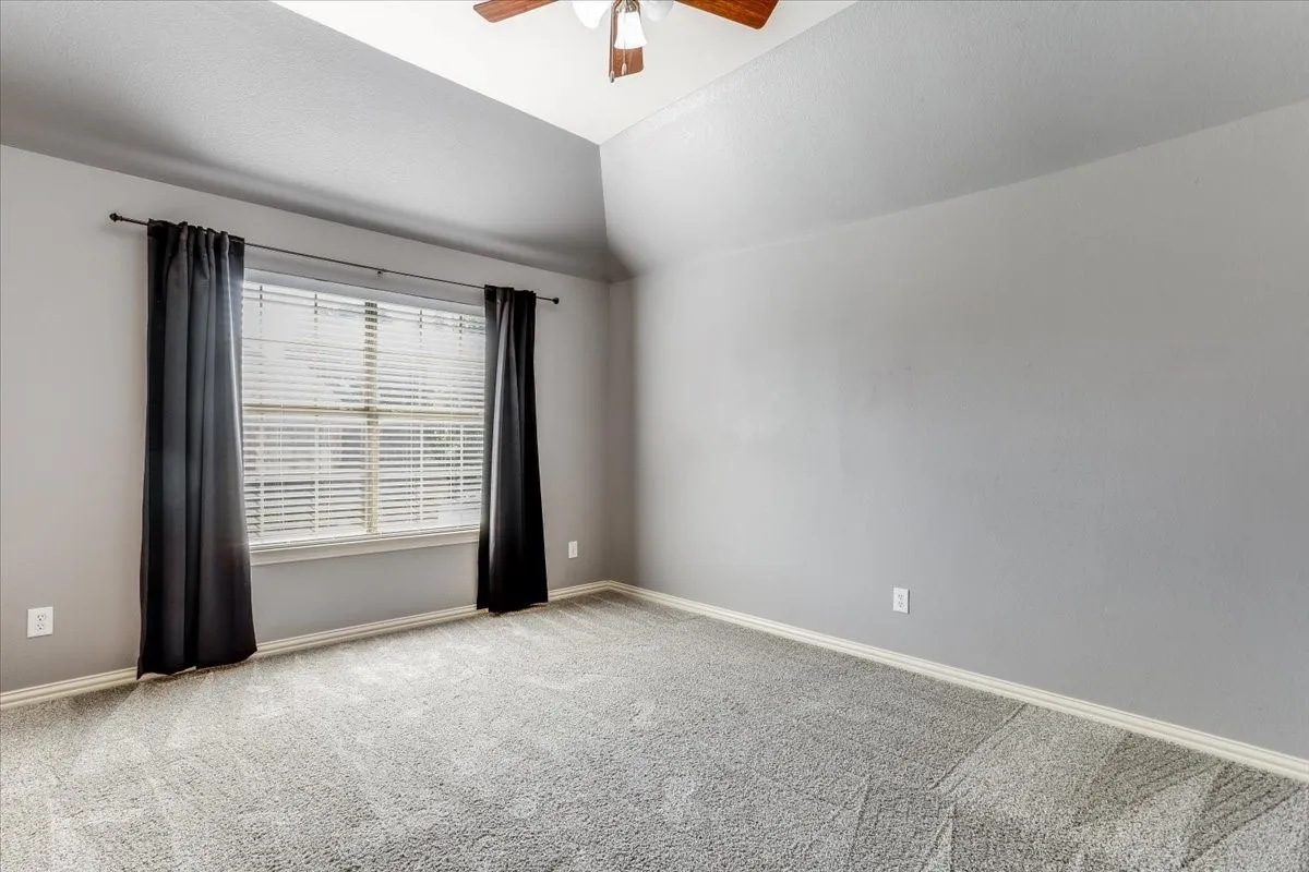Vaulted ceiling, ceiling fan with light fixture, window with blinds, wall-to-wall carpeting, and neutral paint
