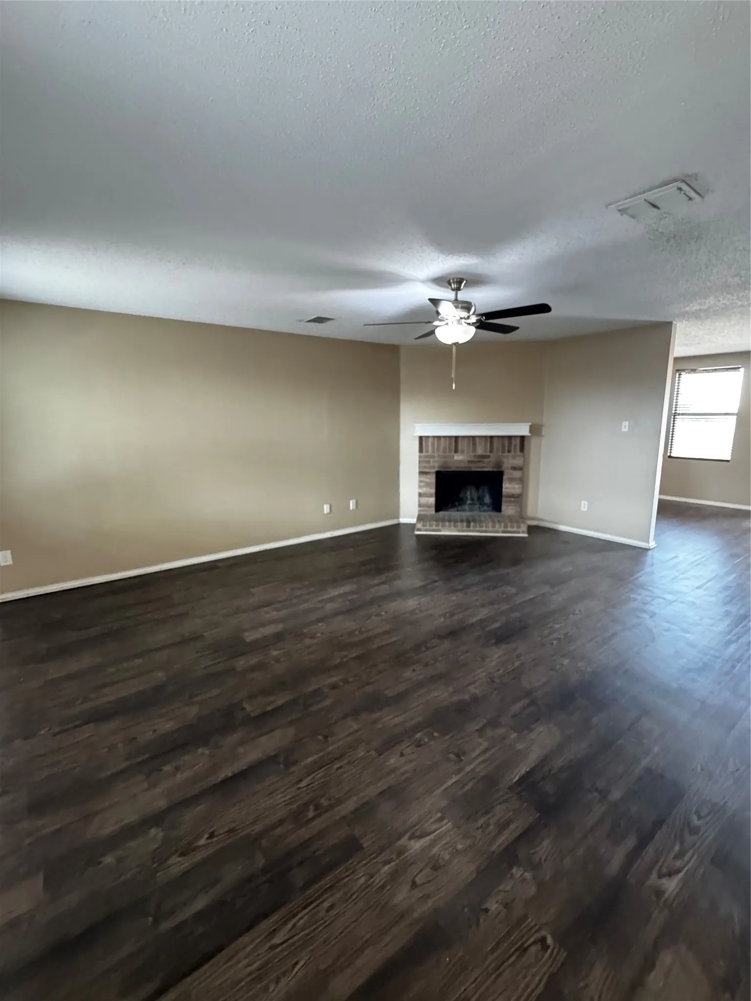 Spacious room featuring wood-finish flooring, a brick-surround fireplace, a ceiling fan with integrated lighting, and neutral wall tones