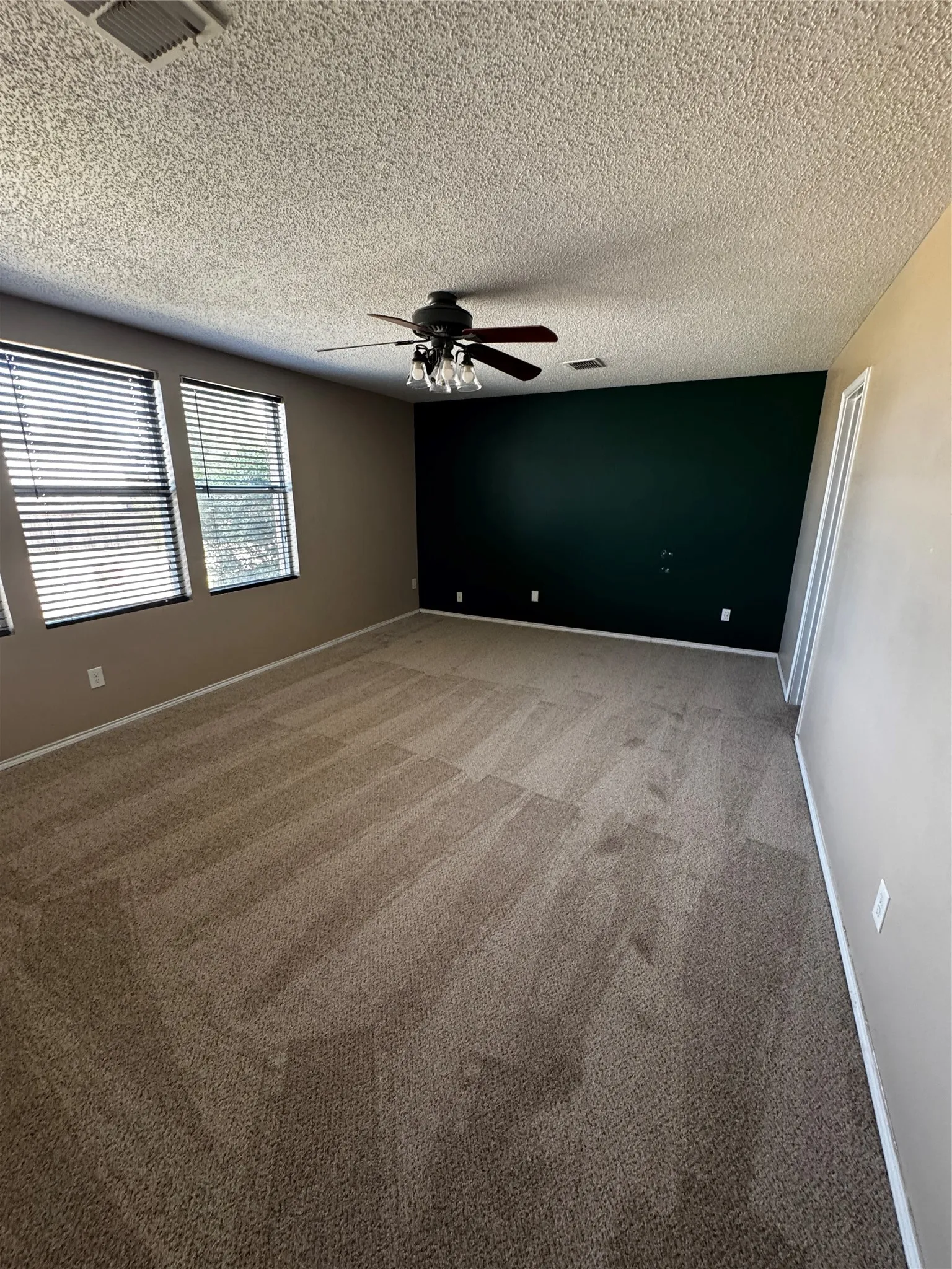 Textured ceiling with a ceiling fan light fixture, two windows with horizontal blinds, neutral-toned walls, and patterned carpet flooring