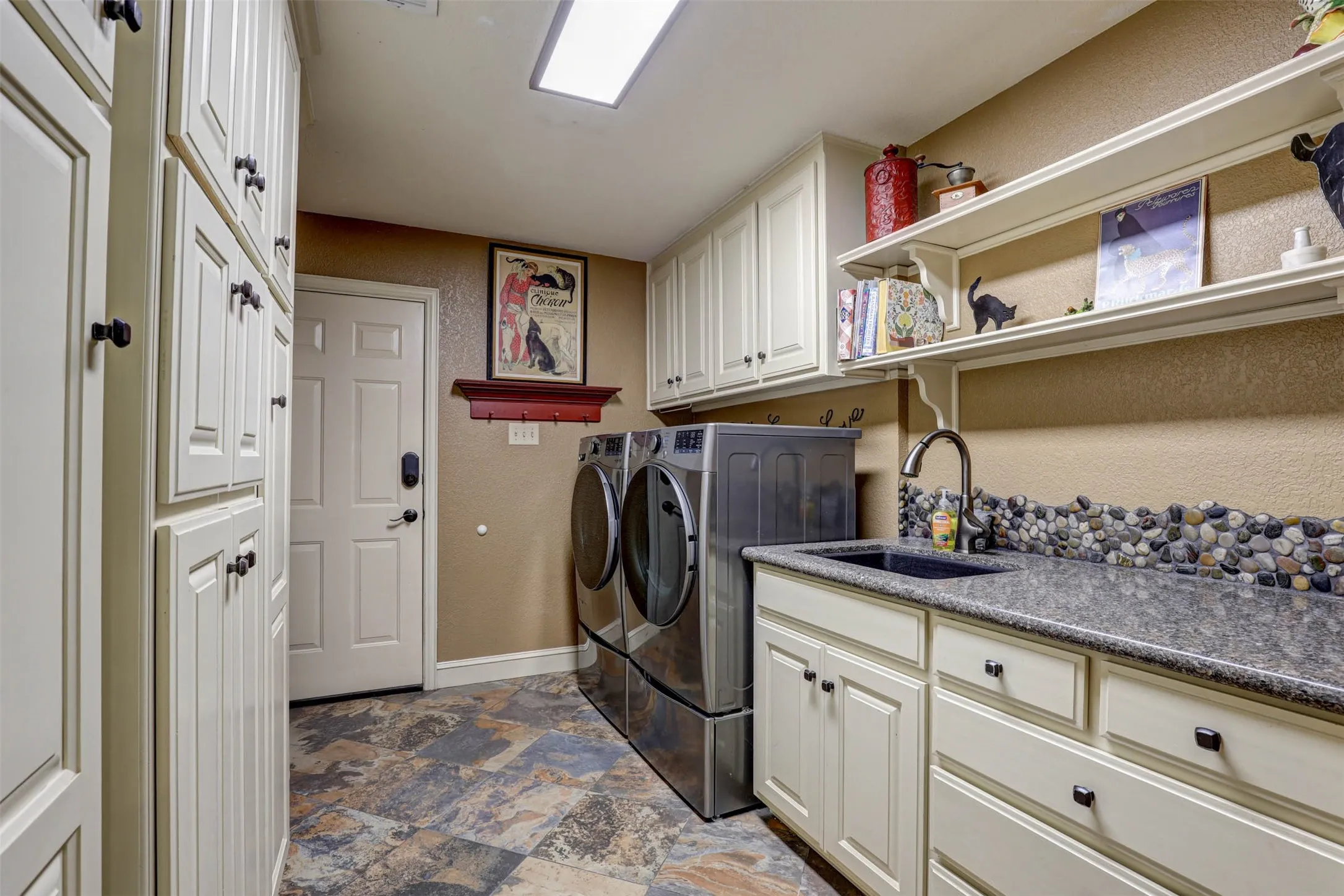 Spacious 14x9 utility room with generous cabinetry, shelving, and a pebble mosaic backsplash at the sink.