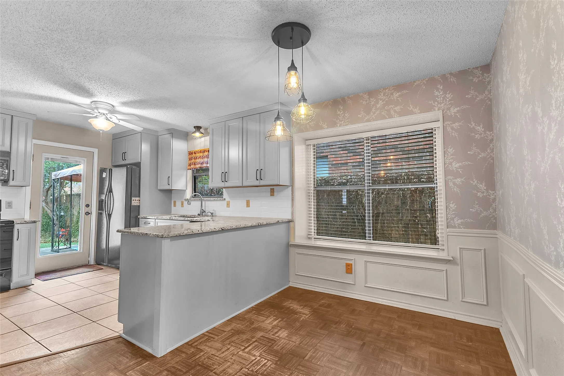 Open-plan kitchen with gray cabinetry, quartz countertops, stainless steel appliances, and pendant lighting over a breakfast bar near a window.
