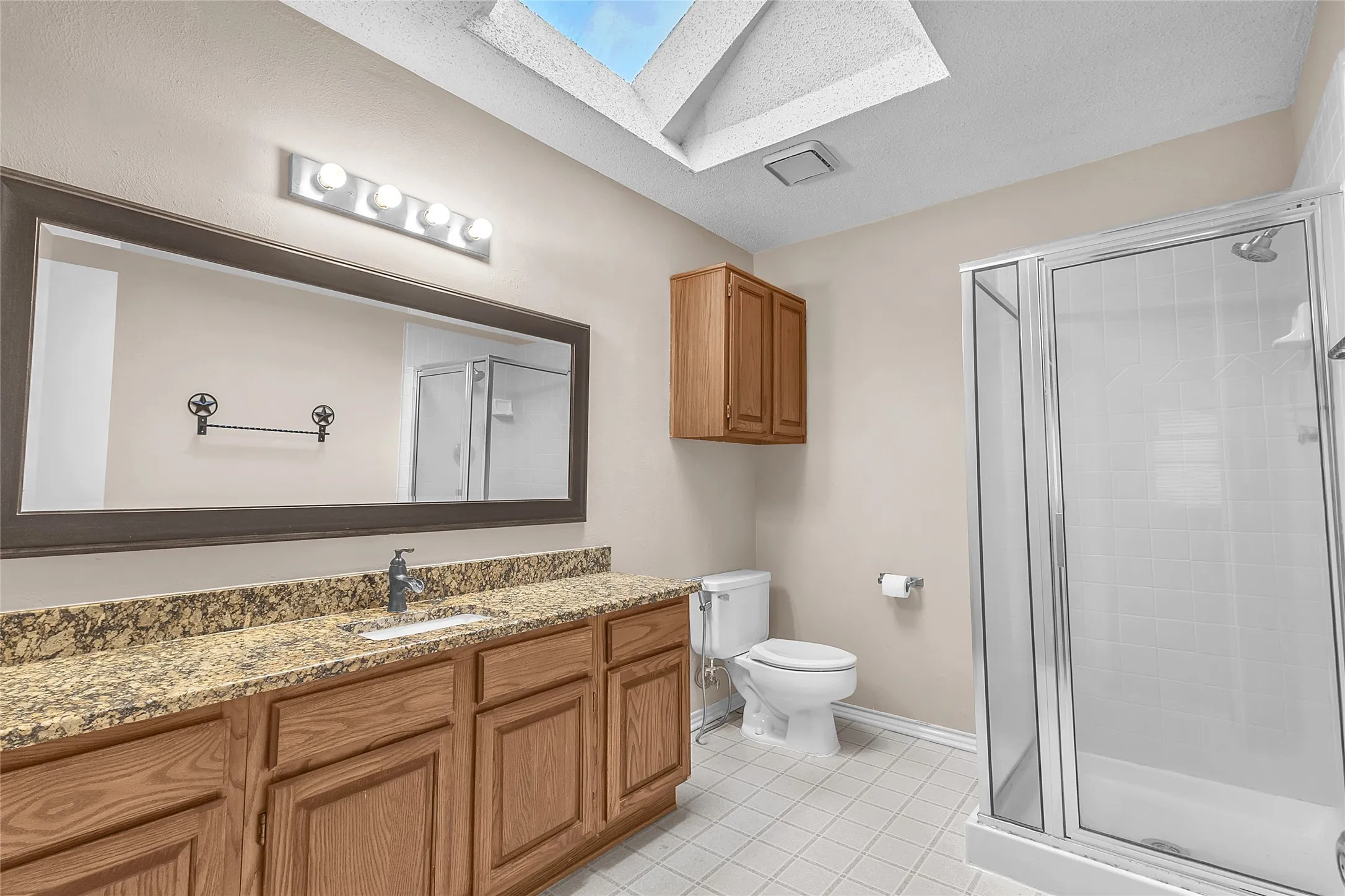 Bathroom with granite countertops, wood cabinetry, porcelain tile flooring, clear glass shower enclosure, and natural light from skylight