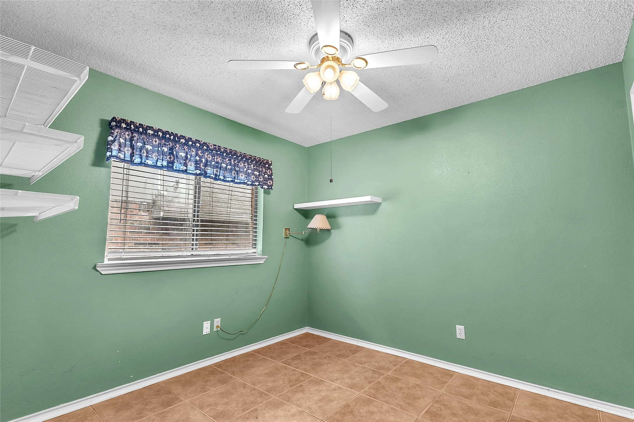 Small green bedroom with tan tile flooring, ceiling fan, window treatments, and wall-mounted shelves.