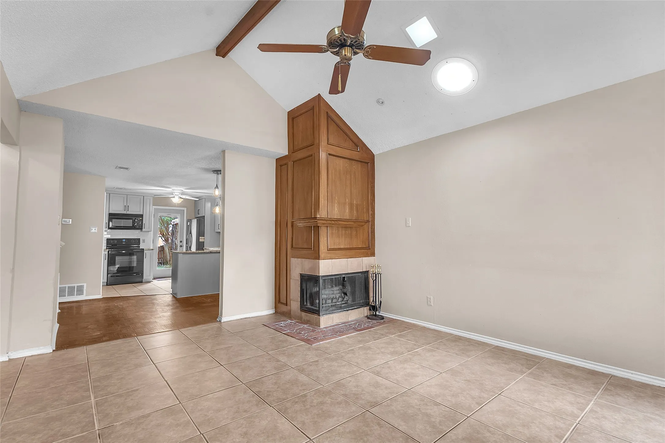 Living room with vaulted ceiling, tile flooring, wooden fireplace surround, and natural light from skylight and side windows.