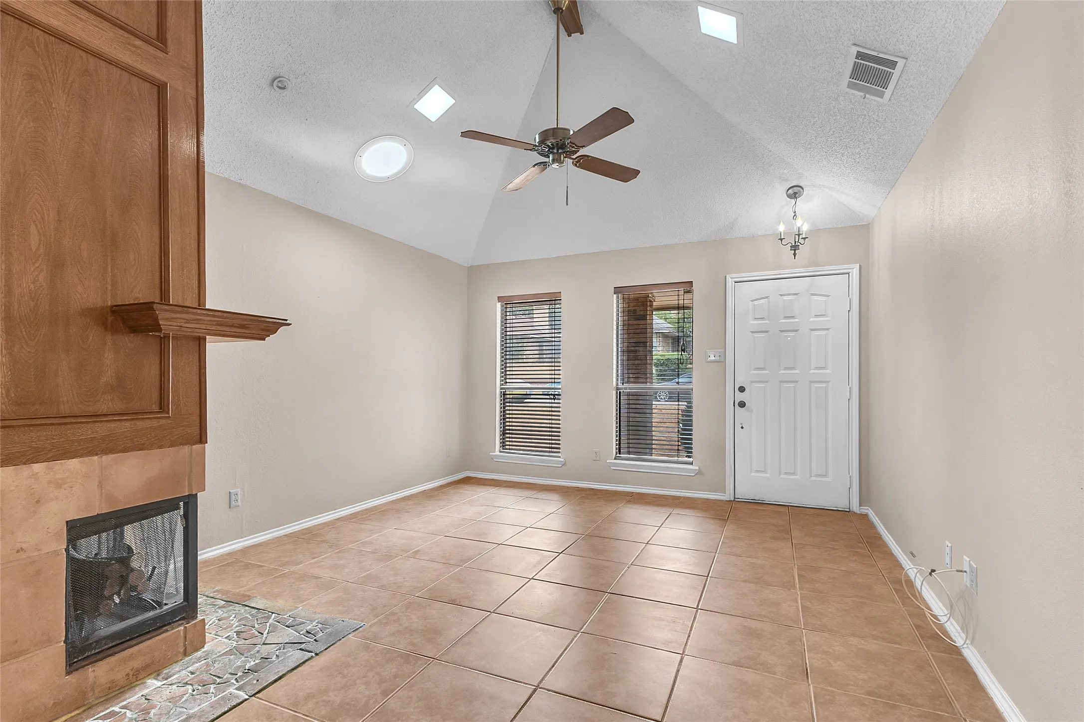Living room with vaulted ceiling, tile flooring, wooden mantel fireplace, and large windows providing natural light.