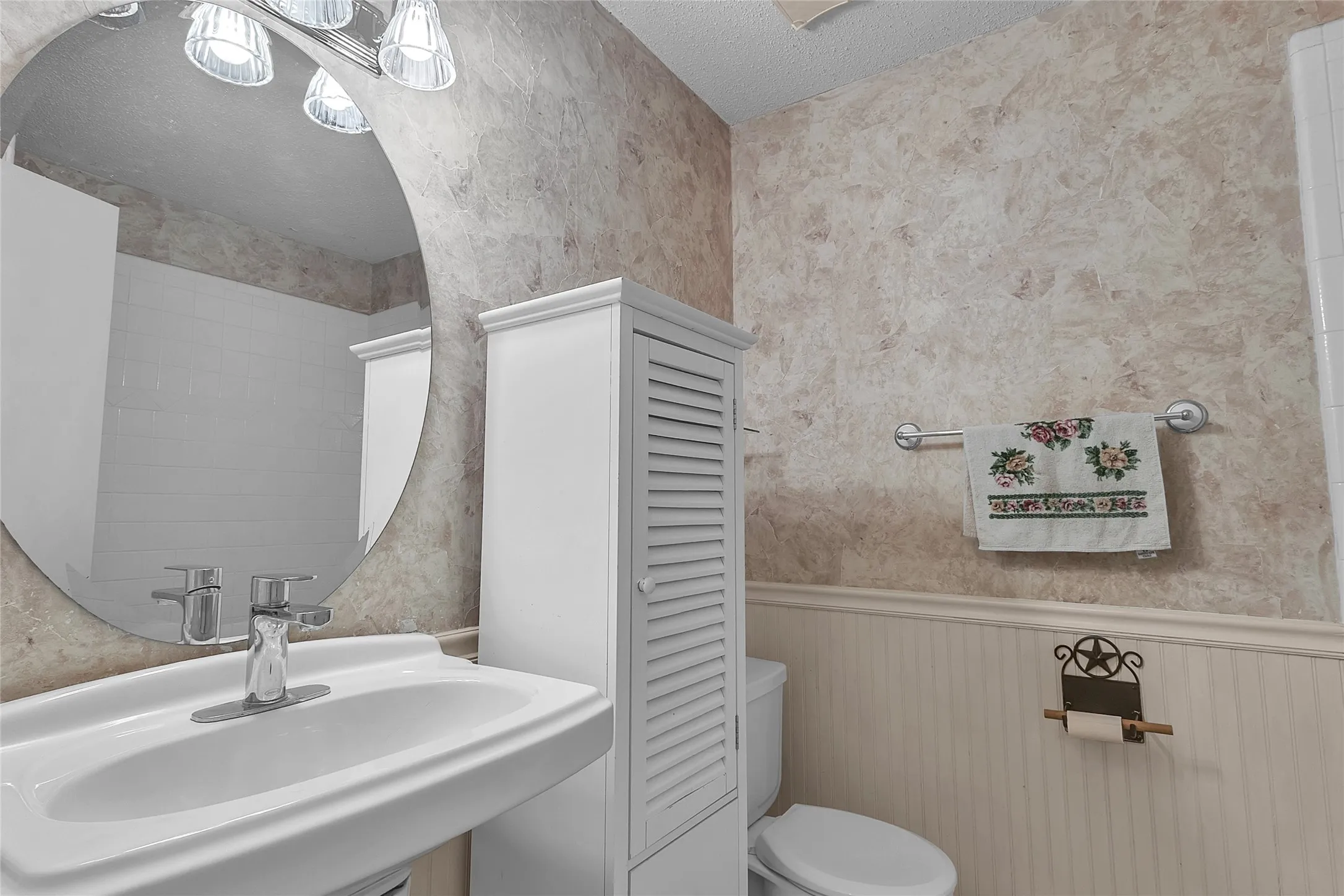 Bathroom with circular mirror, white cabinetry, porcelain sink, and light-colored textured wall finish. Compact layout with a towel rack and recessed lighting.