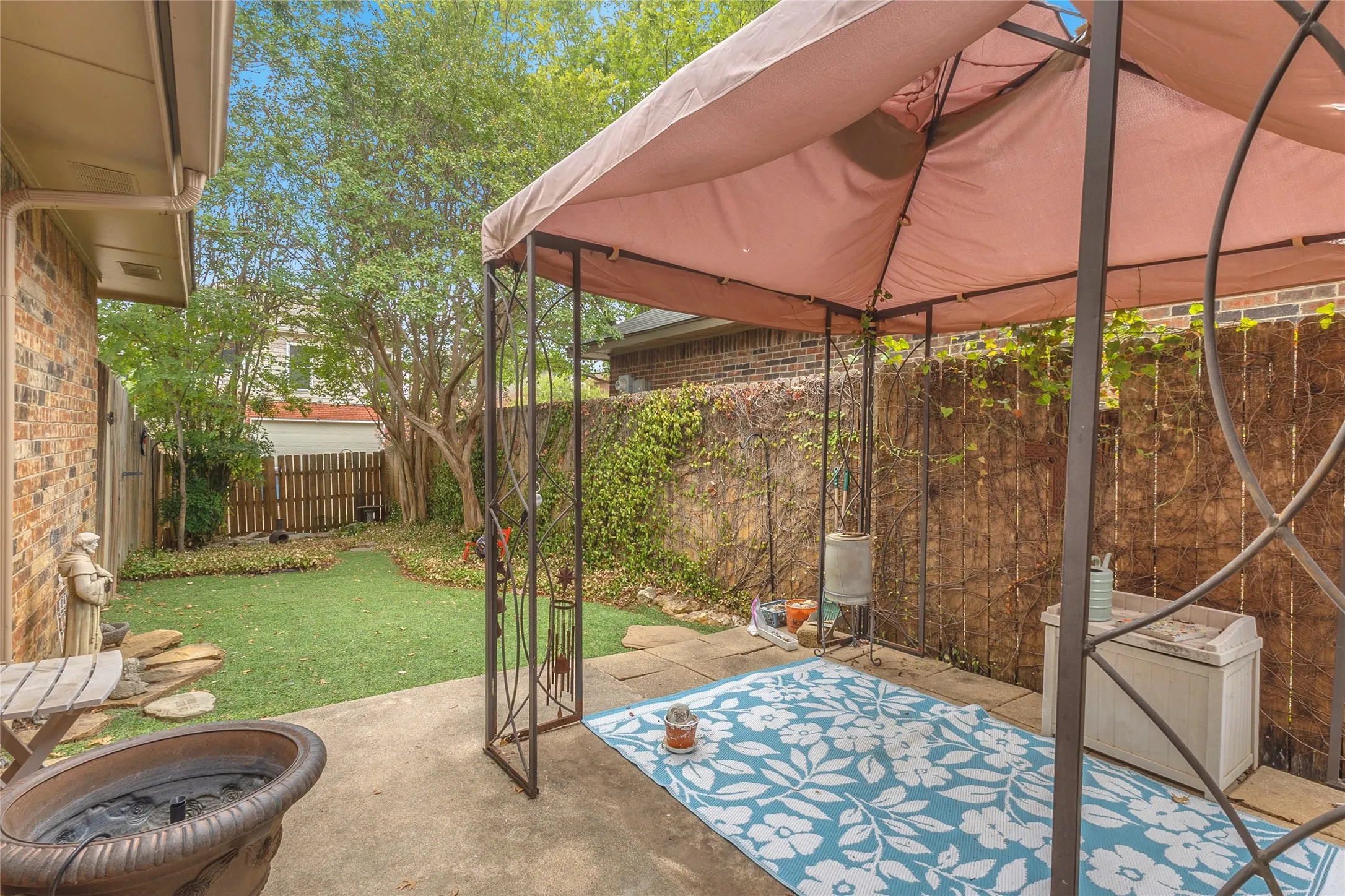Covered back patio with a light pink canopy, stone pavers, artificial grass, and climbing vines along the wall for a private outdoor space