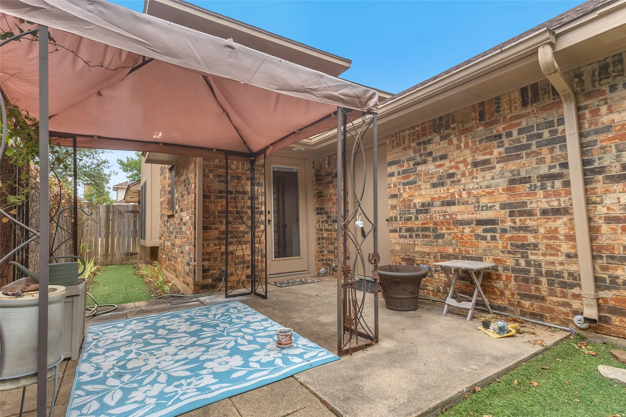 Covered back patio with a canvas canopy, brick wall, concrete flooring, and a decorative area rug. Outdoor space with potential for relaxation.