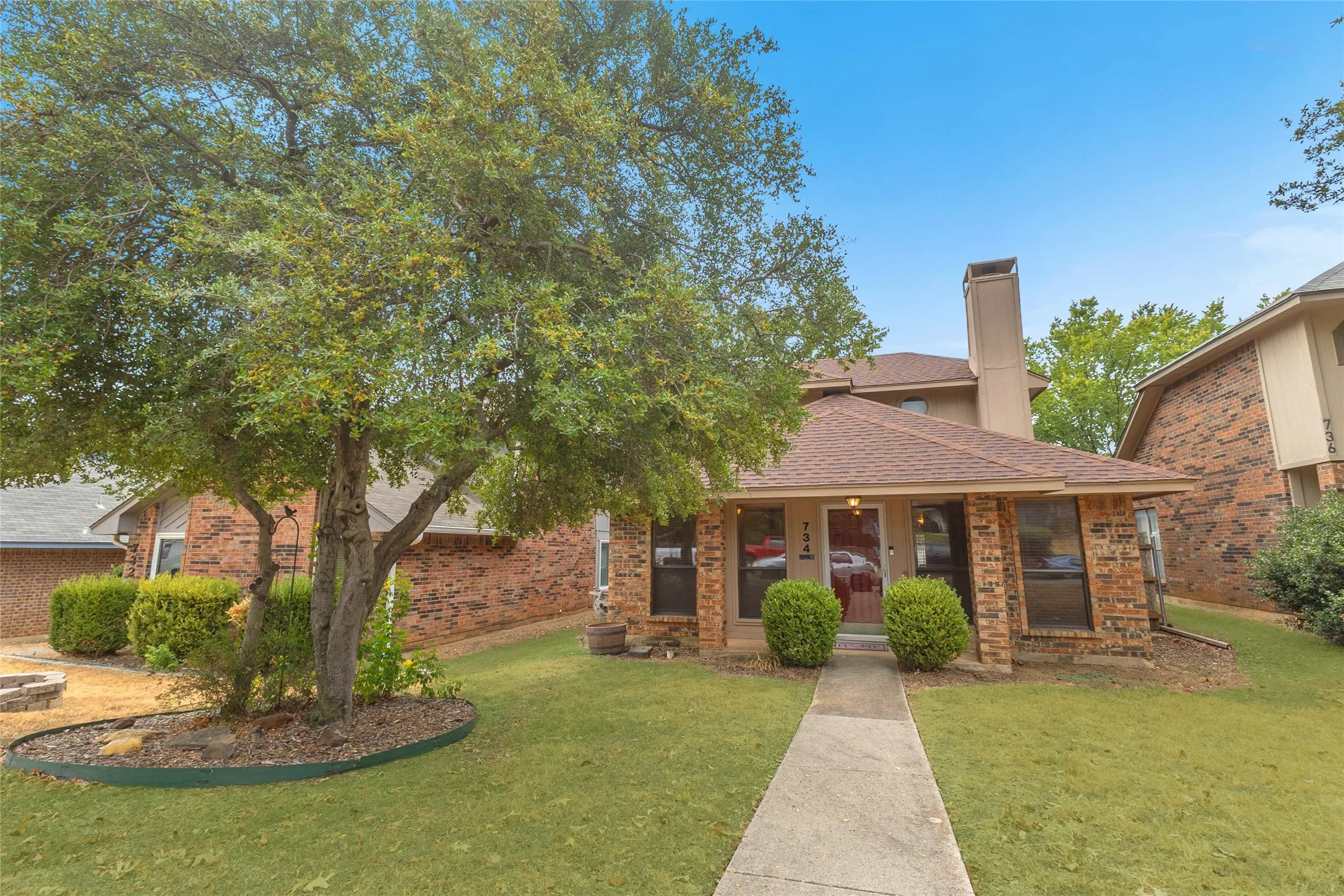Front porch with brick façade, light landscaping, and shaded by a mature tree in a residential neighborhood with a clear blue sky.