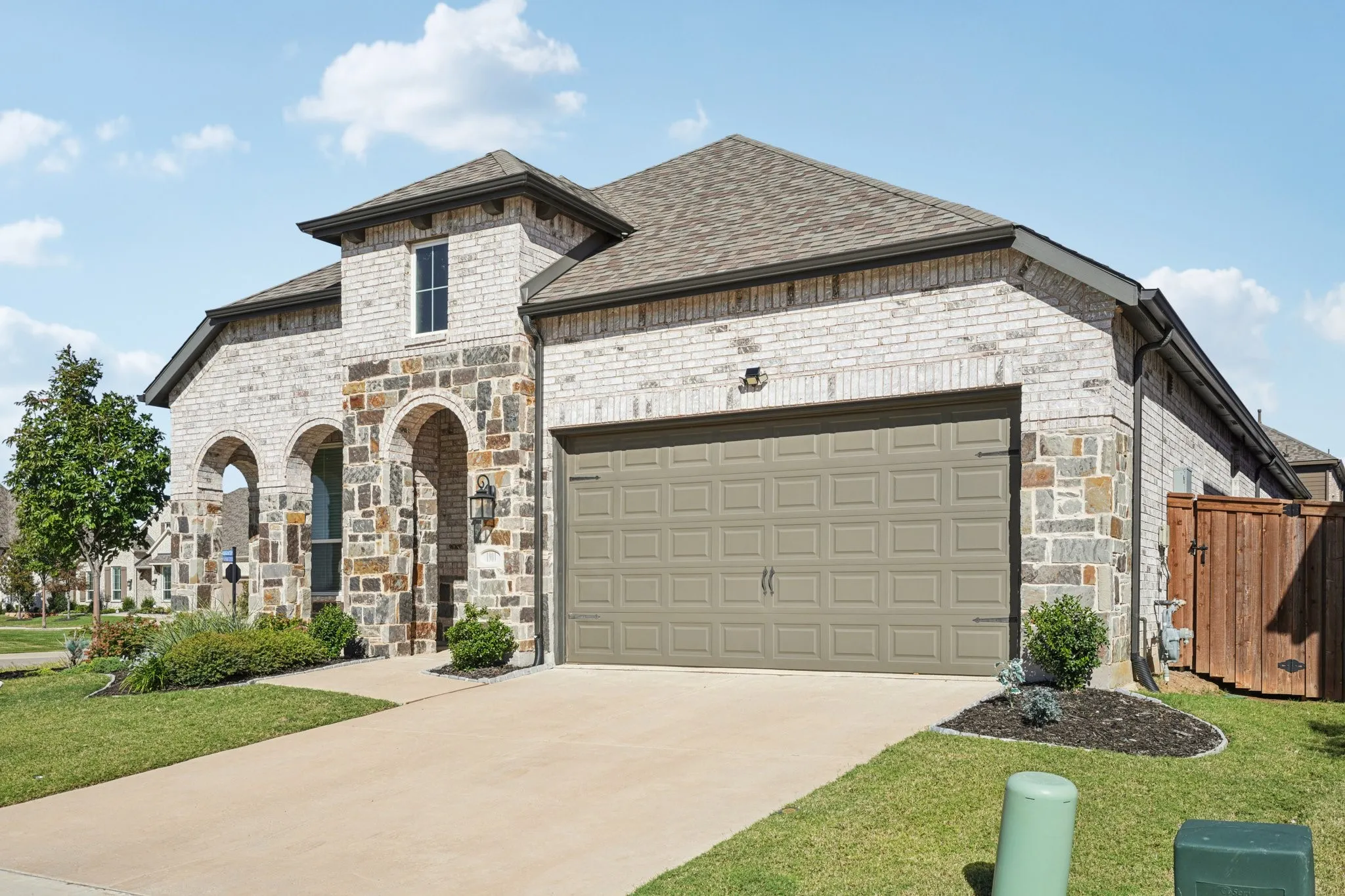 Multi-textured brick and stone exterior with arched entryways, an attached garage, paved driveway, and established landscaping