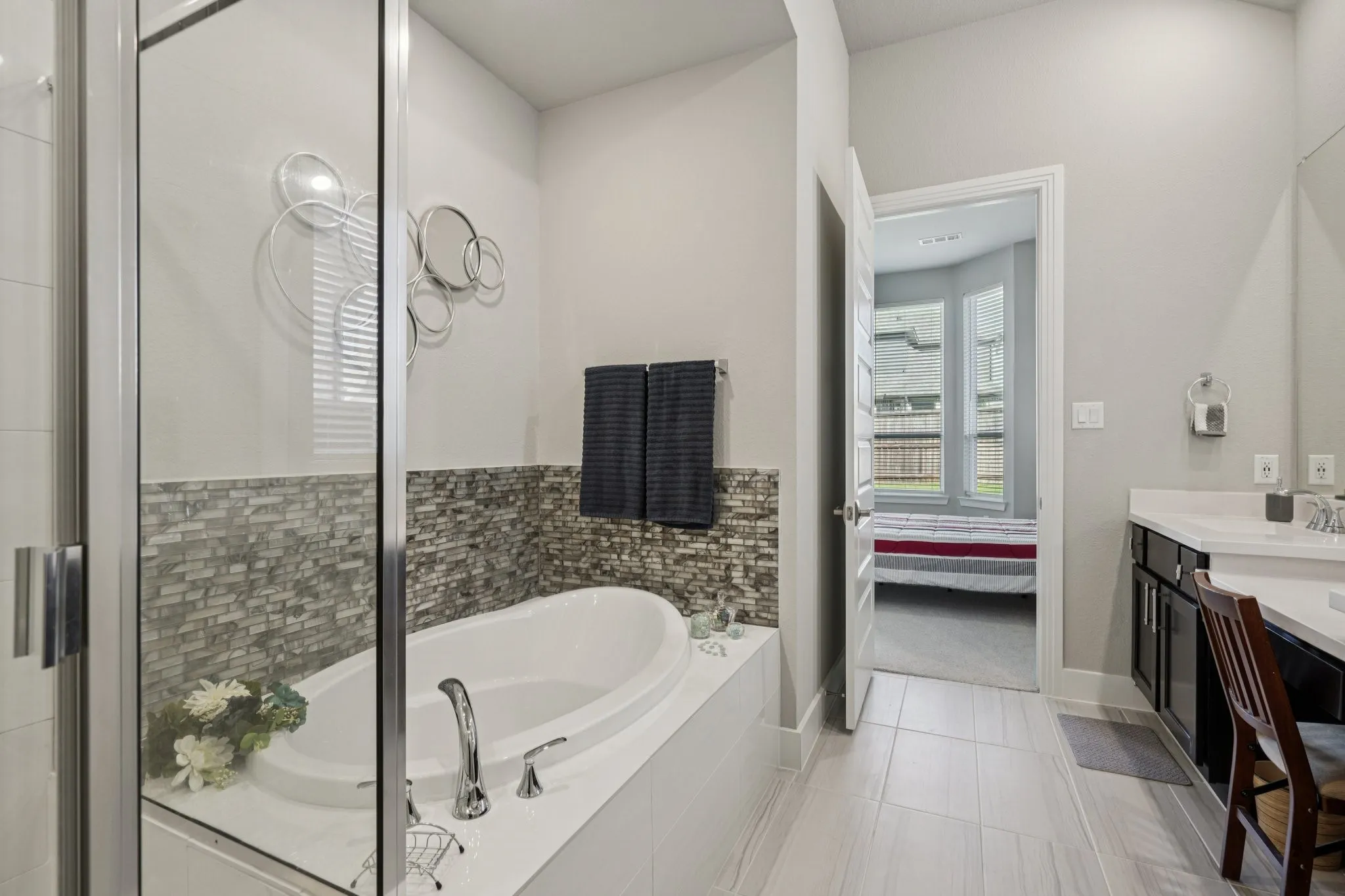 Built-in soaking tub with chrome fixtures, glass-enclosed shower, wall-mounted vanity with integrated sink, wood-finish flooring, and a decorative tile accent wall