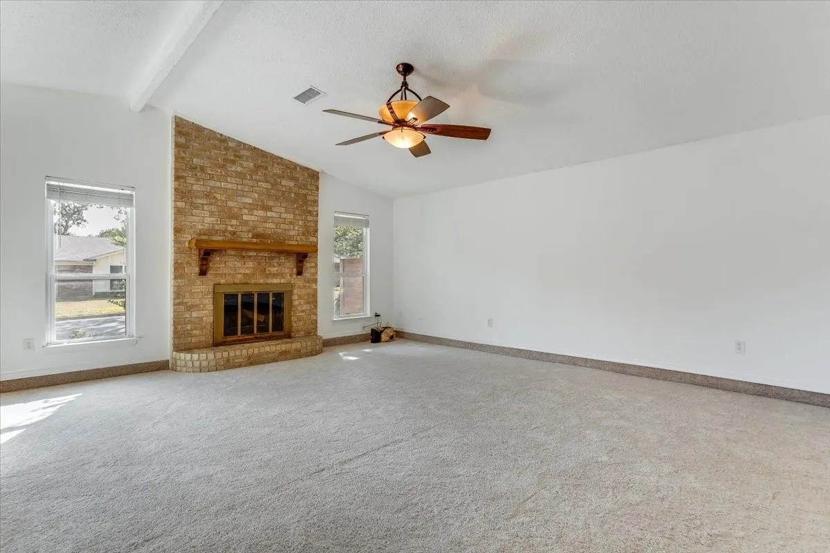 Vaulted ceiling with exposed beam, floor-to-ceiling brick fireplace with wood mantel, ceiling fan with integrated lighting, and dual windows