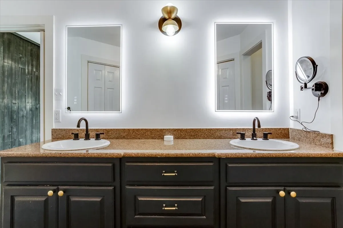 Dual vanity with a stone-finish countertop, undermount sinks, dark wood cabinetry, and backlit mirrors