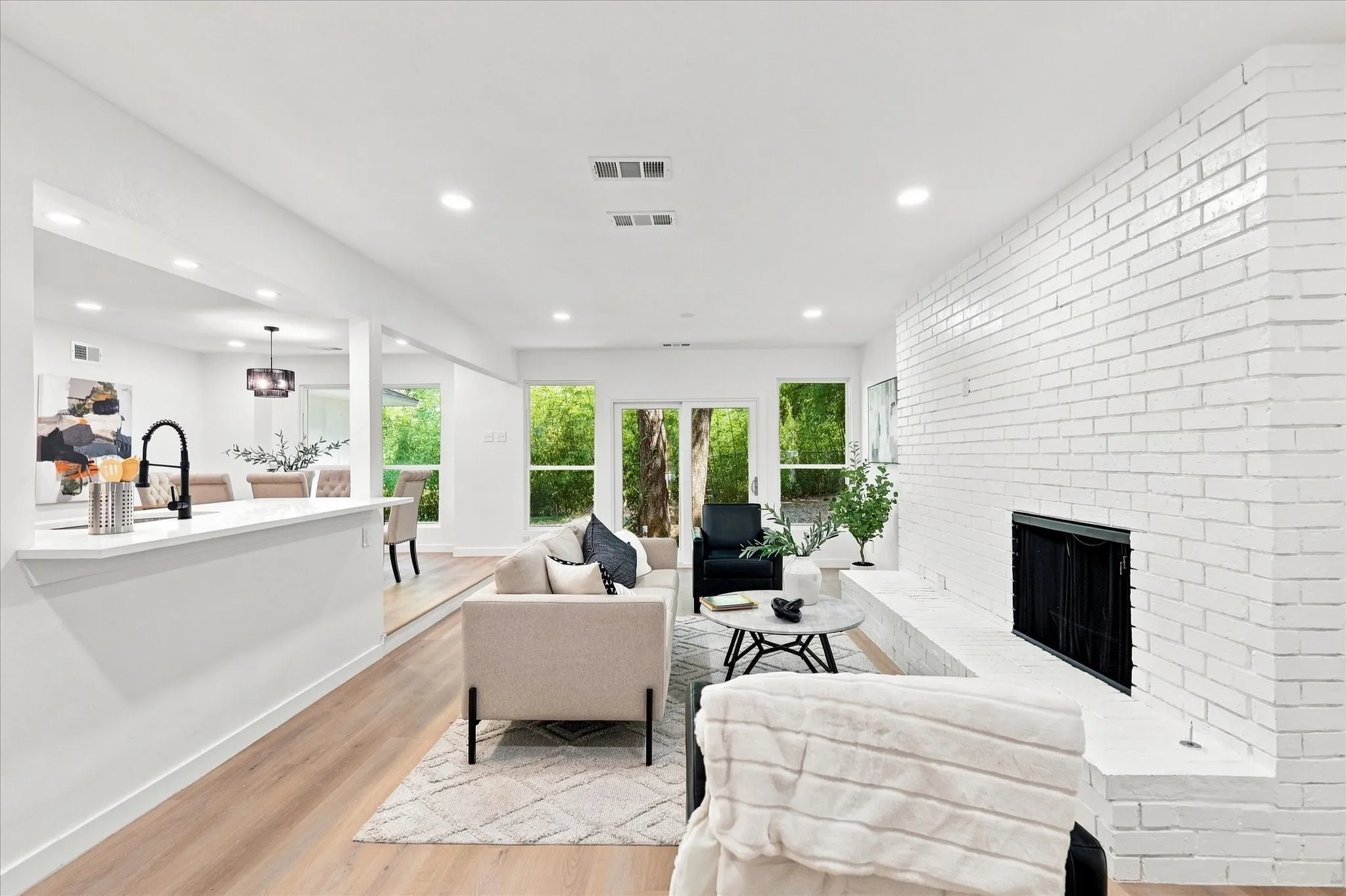 Recessed lighting illuminates the living space featuring a white brick fireplace with a hearth, wood-finish flooring, and large windows