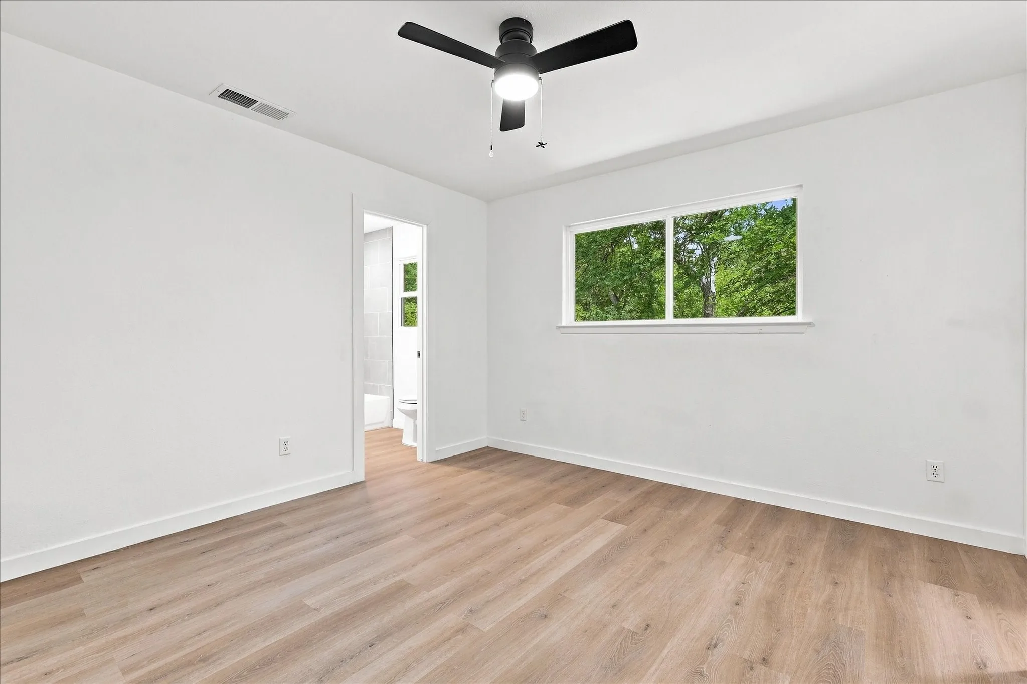 Wood-finish flooring, white walls, ceiling fan, and a window framing a verdant outdoor landscape