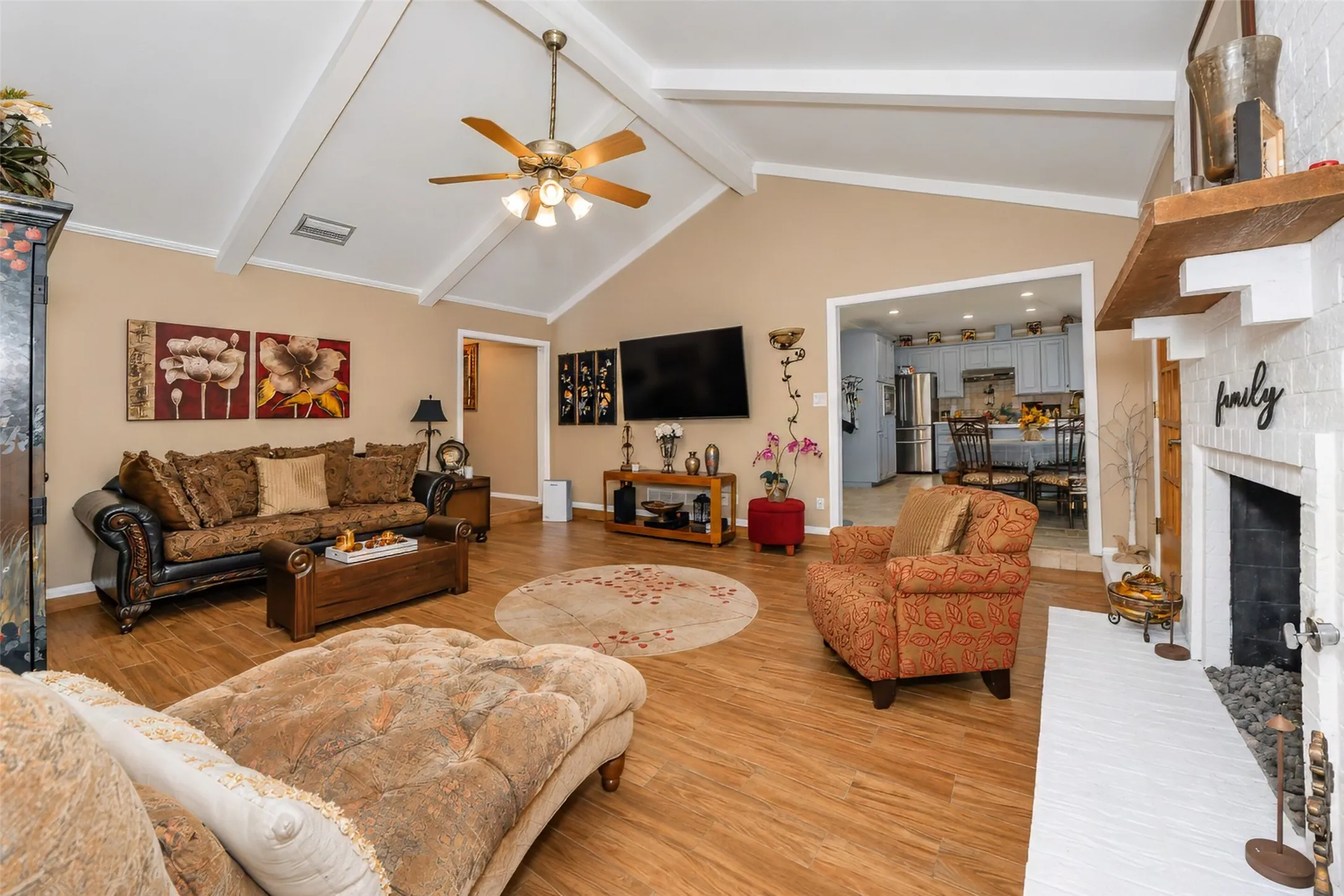 Vaulted ceiling with exposed beams, wood-finish flooring, white brick fireplace, and an open doorway to a kitchen