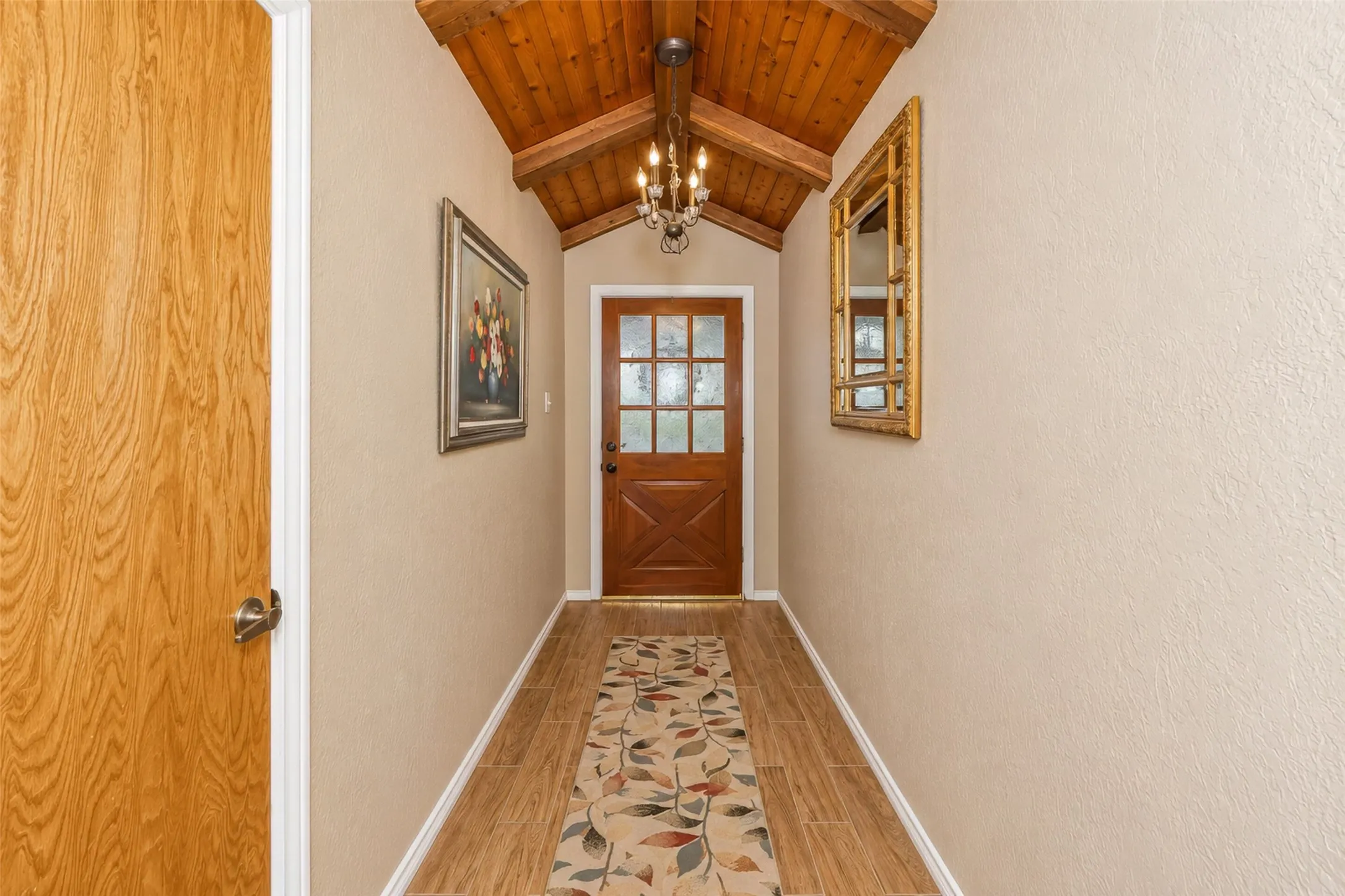 Vaulted wood-paneled ceiling, exposed wood beam, wood-finish flooring, wood entry door with glass panes, and a chandelier