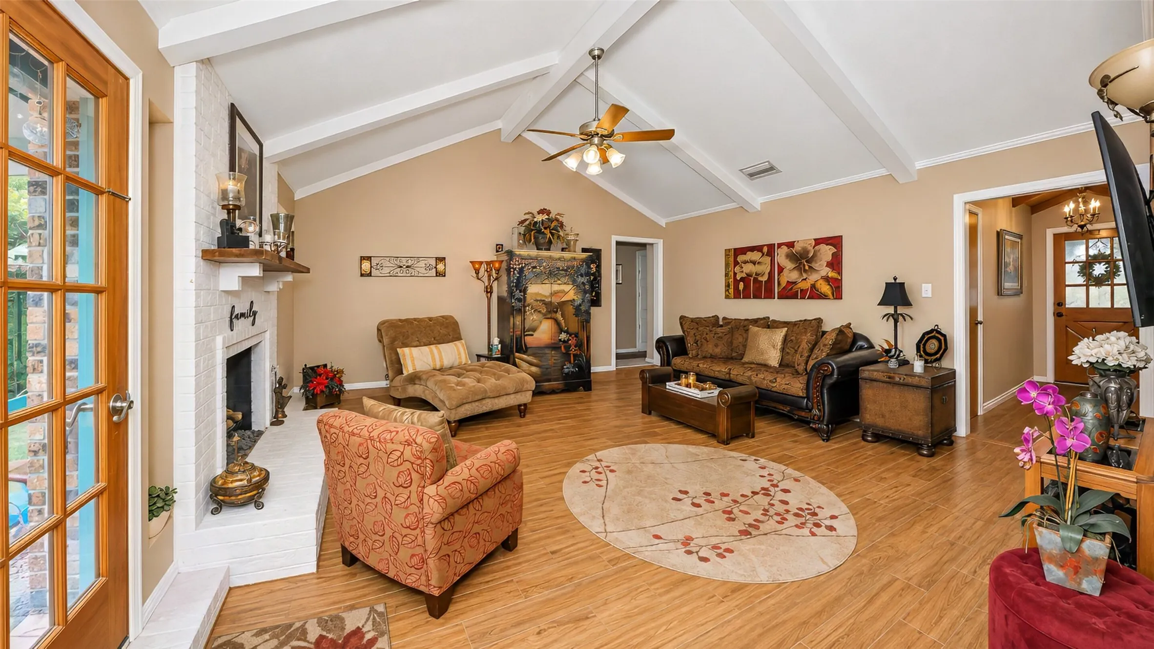 Living area featuring a vaulted ceiling with exposed beams, wood-finish flooring, a white brick fireplace with a wood mantel, a ceiling fan, and French doors
