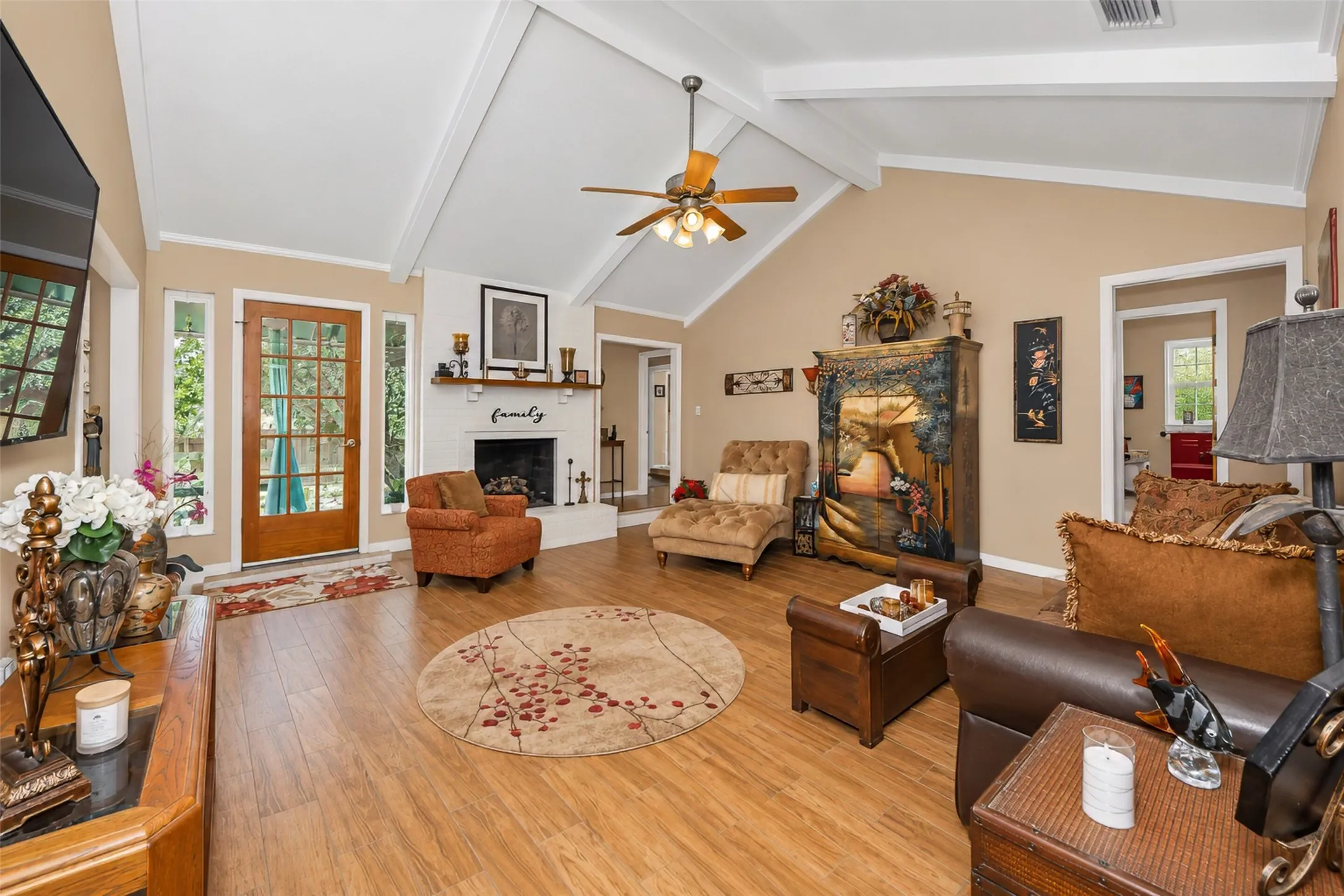 Vaulted ceiling with exposed beams, wood-finish flooring, white brick fireplace, ceiling fan, and a wood-paneled door with sidelights