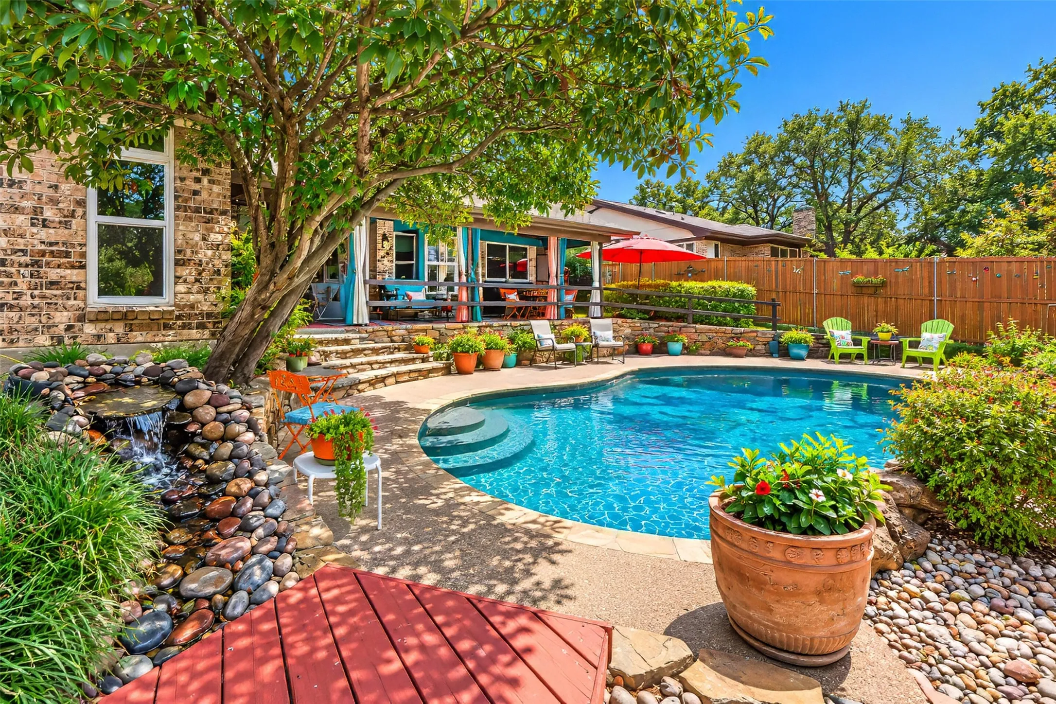 Curved swimming pool with integrated steps, stone waterfall feature, brick exterior, and a wood privacy fence
