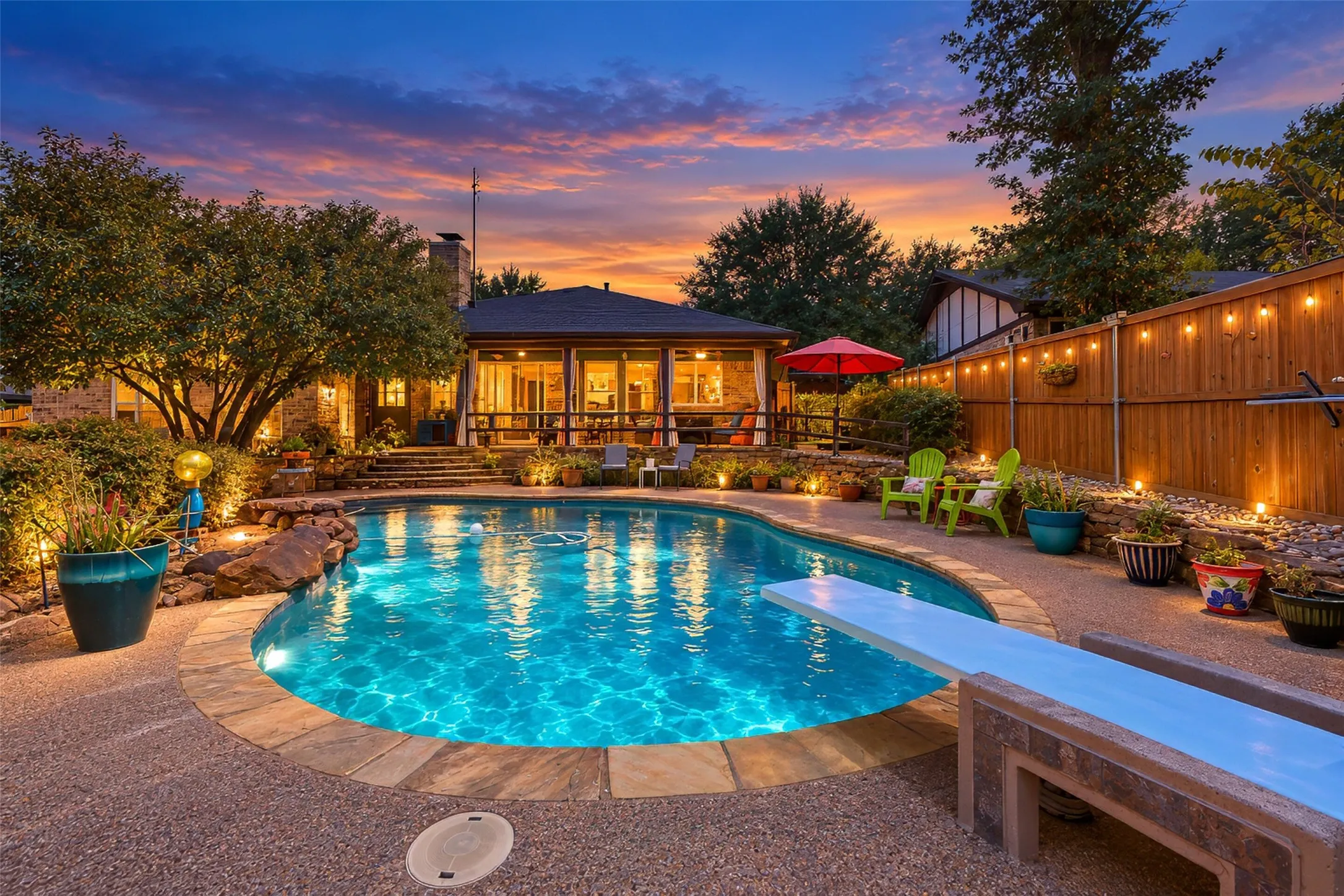 Curved swimming pool with stone coping, a diving board, perimeter landscaping, and a wood privacy fence with string lighting