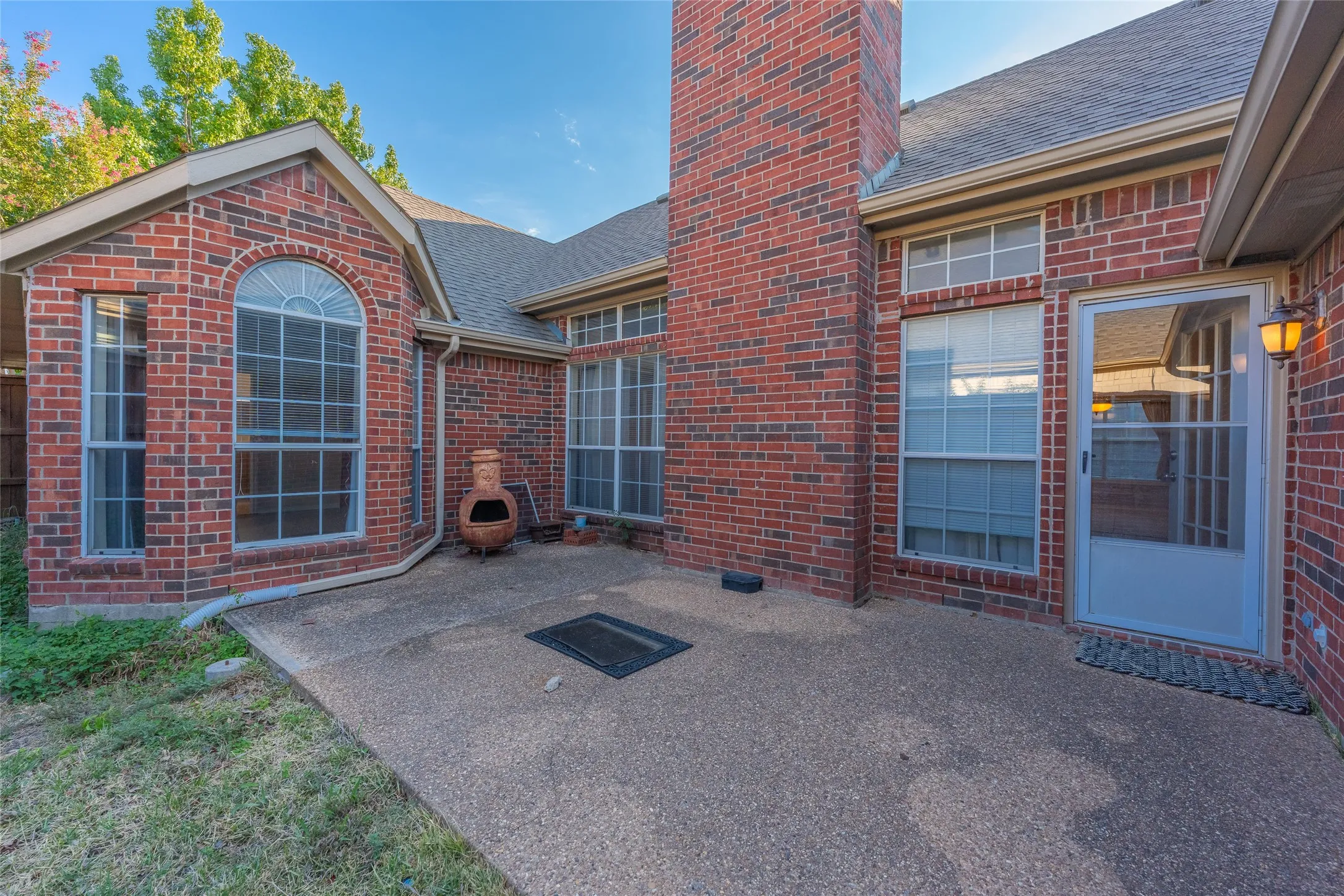 Exterior brick facade with a built-in chimney, arched window, multiple large windows, and a paved patio area