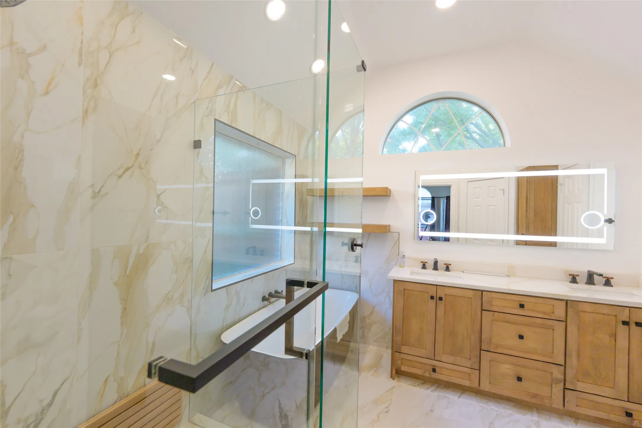 Glass-enclosed shower with a built-in bench, dual vanity with undermount sinks, illuminated mirror, and an arched transom window