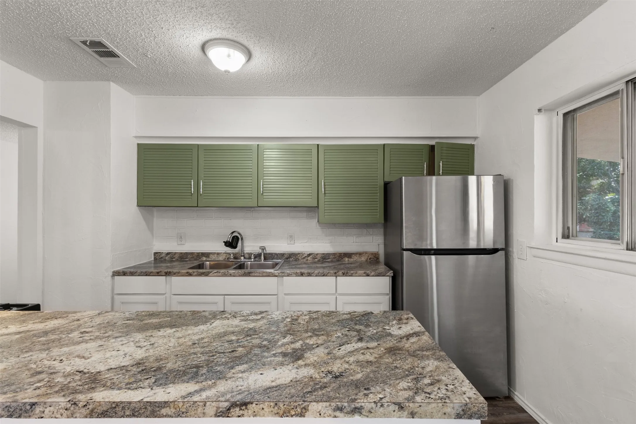 Kitchen featuring stone countertops, a double basin sink, green louvered upper cabinetry, white lower cabinetry, and a stainless steel refrigerator