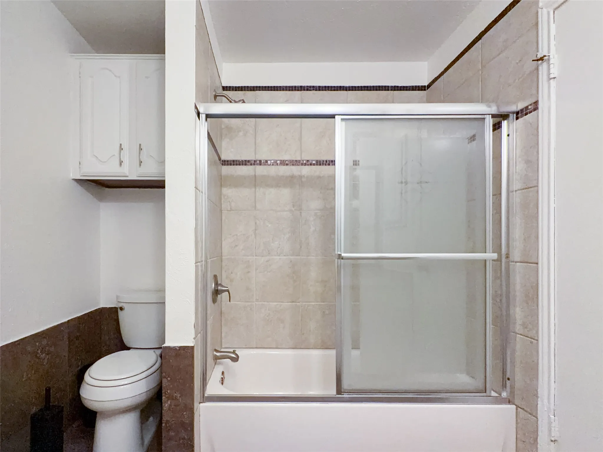 Bathtub with sliding glass enclosure, light-toned wall tiling with decorative accent band, built-in white cabinetry, and toilet with dark-toned wainscoting