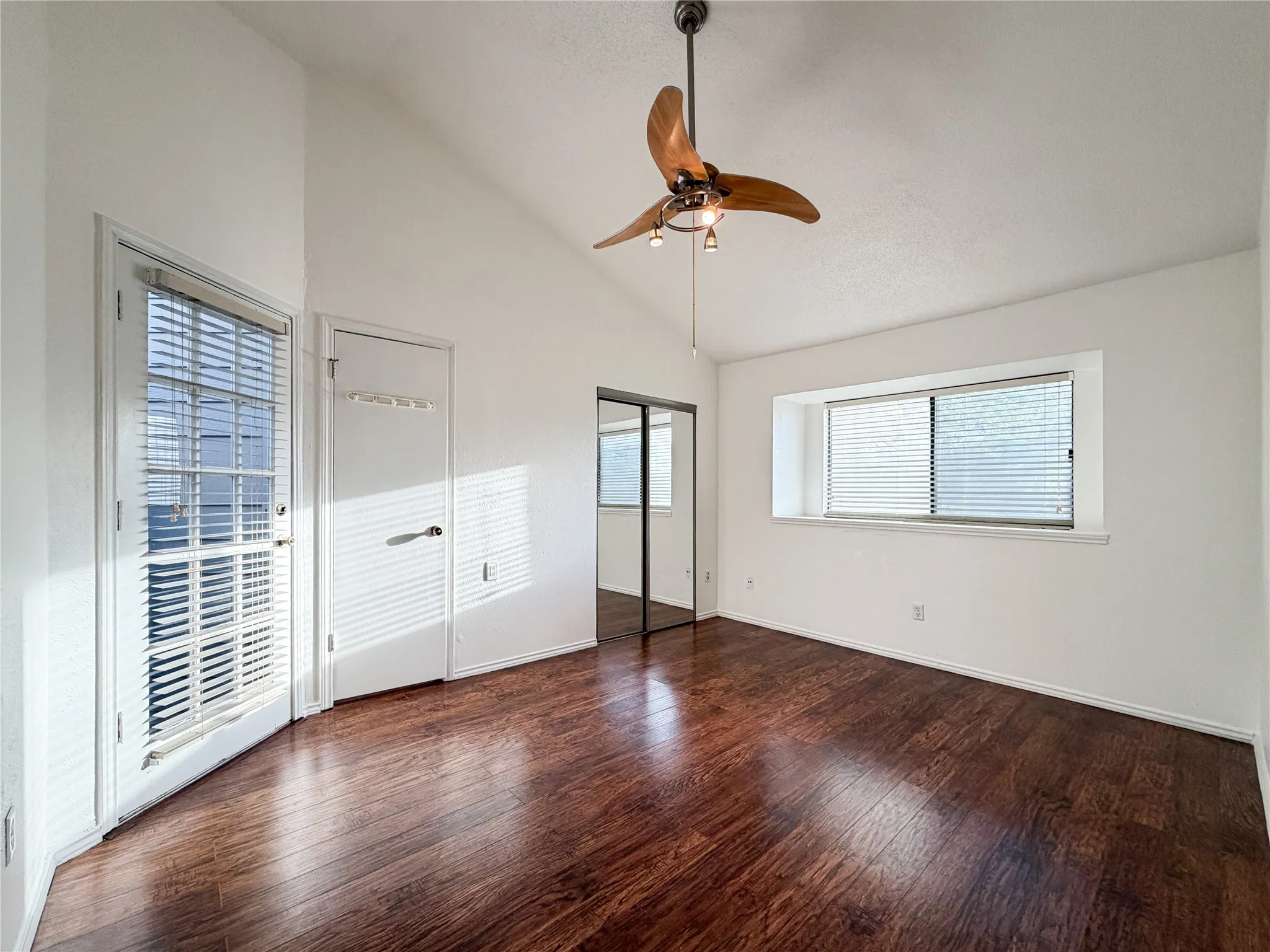 Vaulted ceiling, wood-finish flooring, ceiling fan with integrated lighting, glass-paneled door, and mirrored sliding closet doors