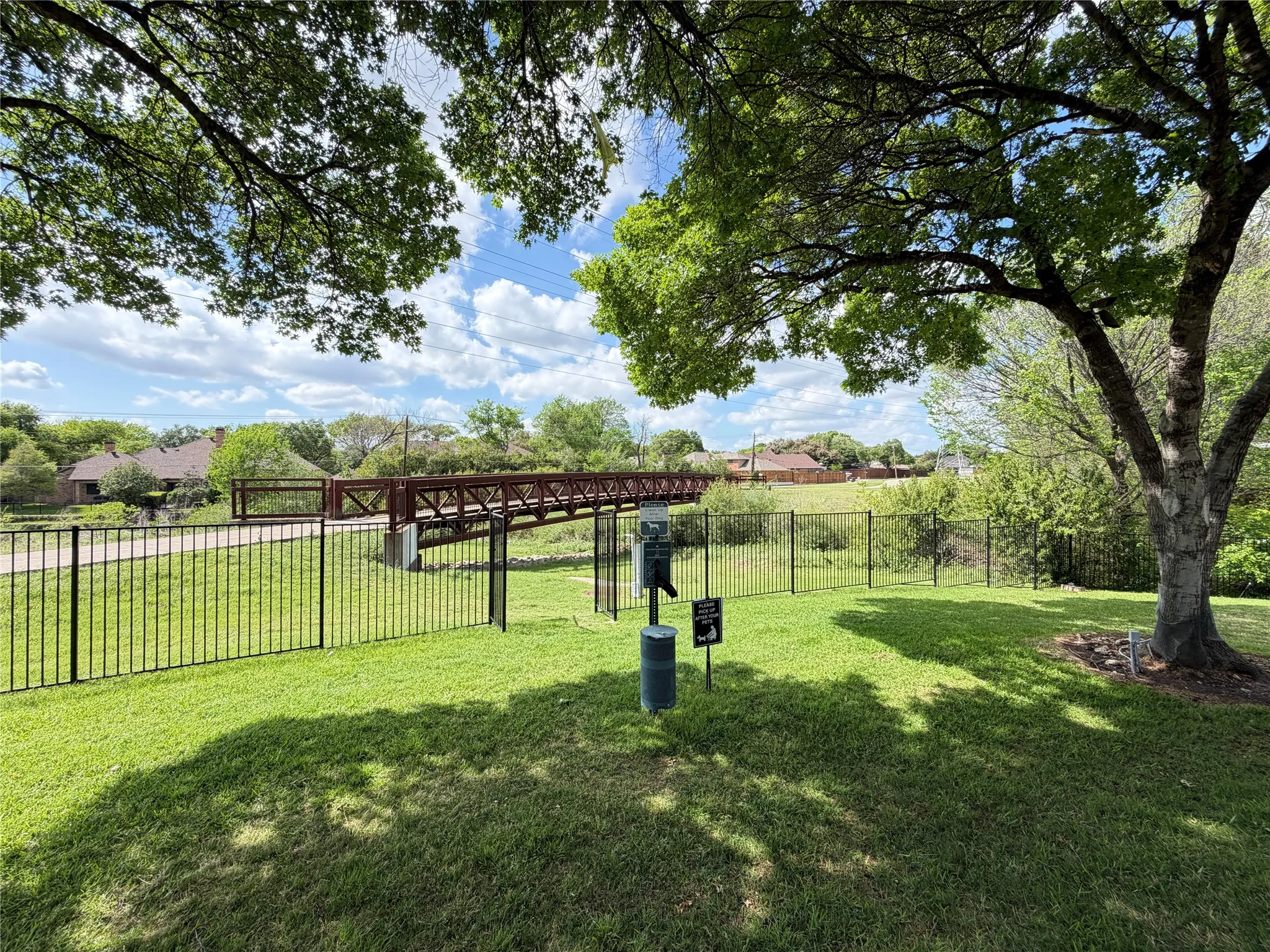 Manicured lawn area with mature trees, featuring a black metal fence and a pedestrian bridge