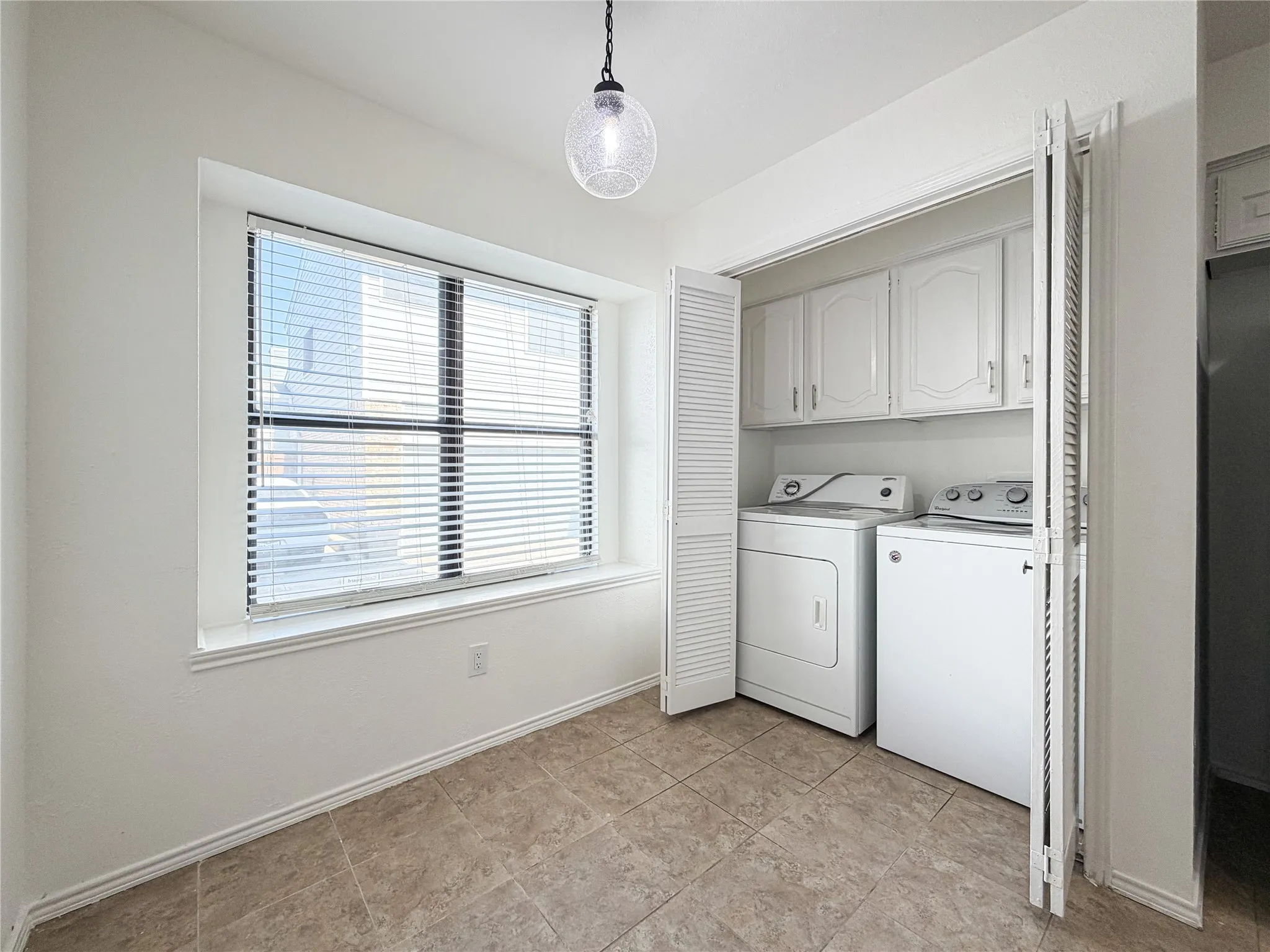 Laundry area featuring neutral-toned tile flooring, a window with blinds, and a built-in closet with louvered bi-fold doors