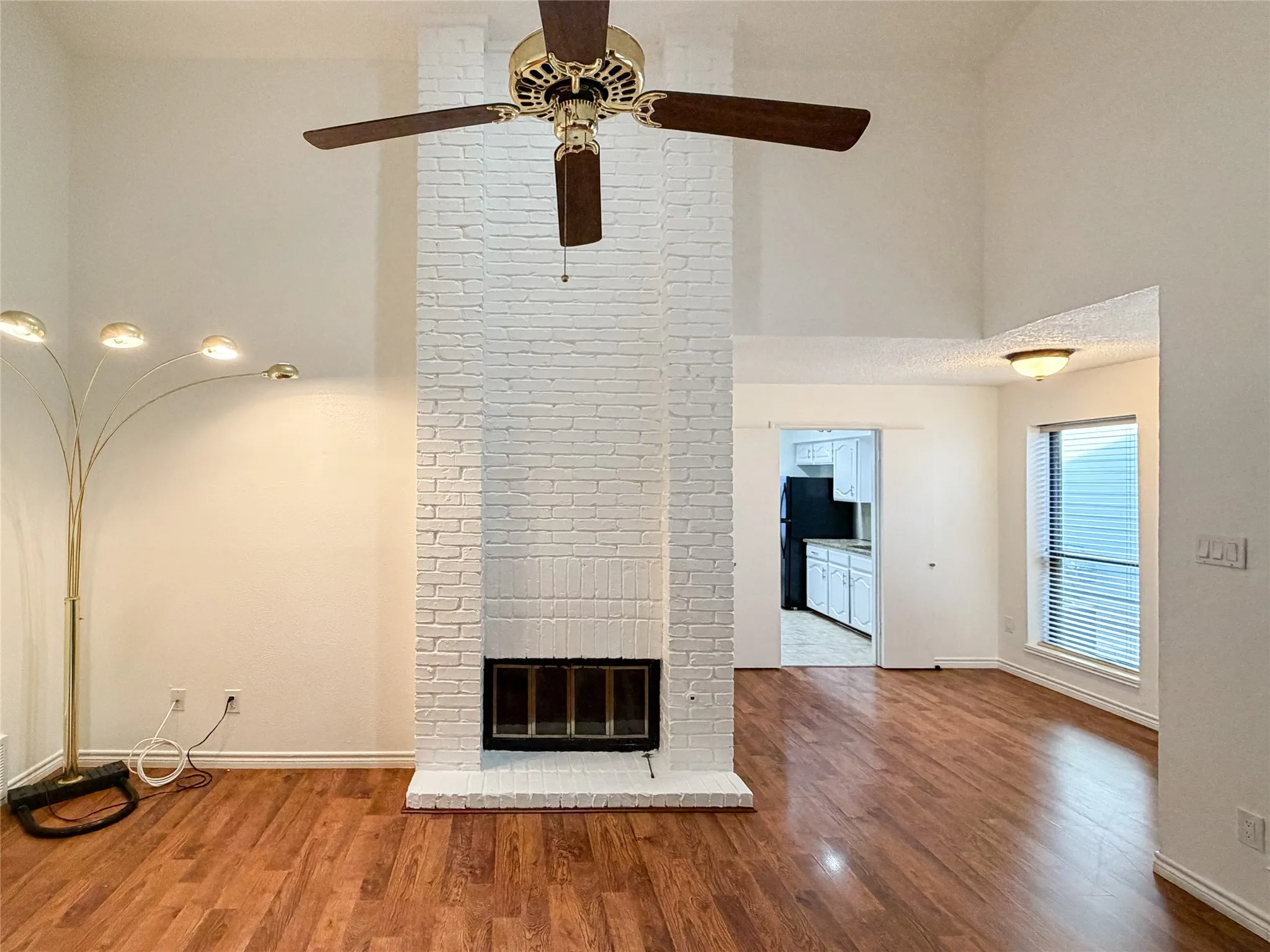 Vaulted ceiling with a ceiling fan, white-painted brick fireplace, wood-finish flooring, and window with blinds