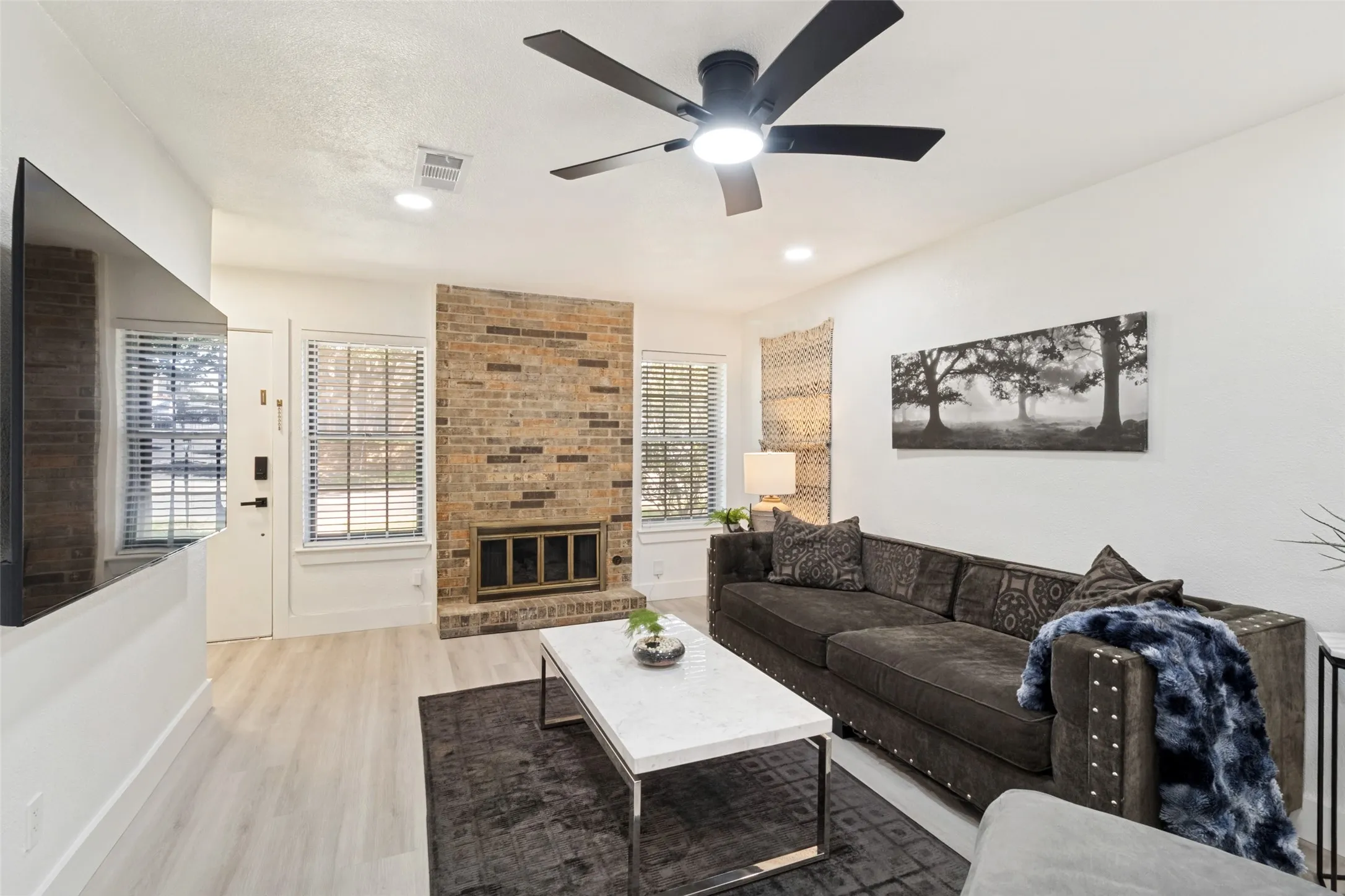 Brick fireplace feature, wood-finish flooring, recessed lighting, ceiling fan with integrated light, and multiple windows with blinds