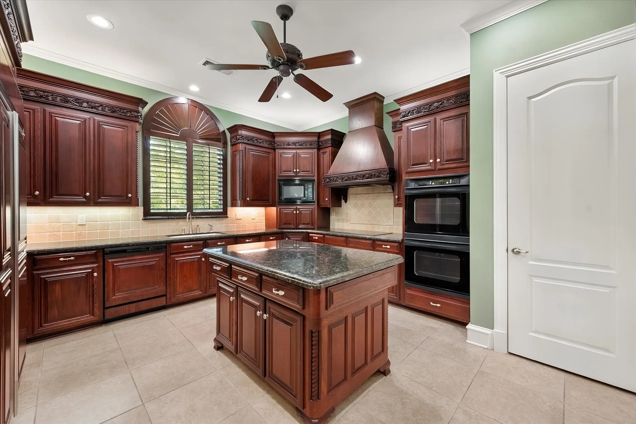Kitchen featuring decorative vent hood and solid wood cabinetry, a central island with a stone countertop, a tile backsplash, and built-in double ovens
