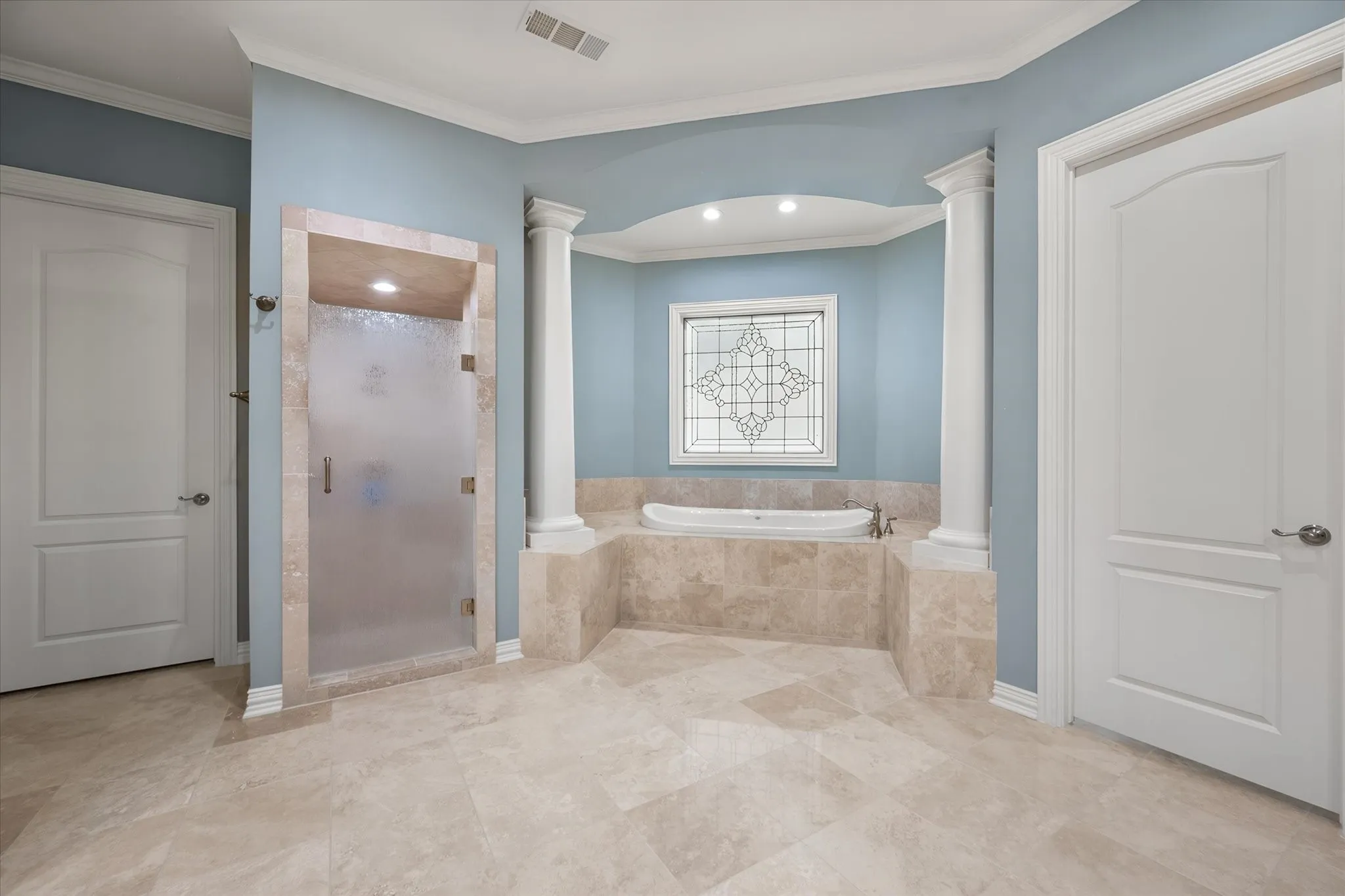 Expansive tile flooring, a large soaking tub with a tiled surround, a separate glass-enclosed shower, decorative columns, and a leaded glass window