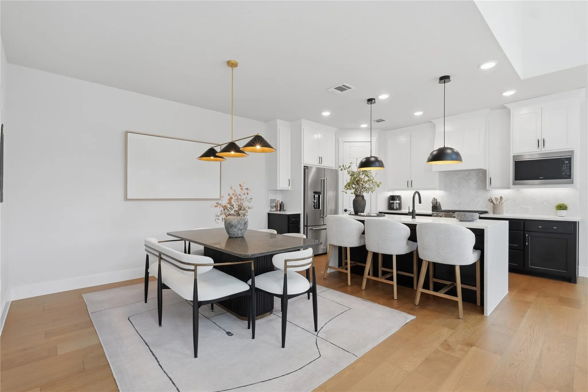 Light wood-finish flooring, white walls, recessed ceiling lights, an island with countertop seating, and a kitchen featuring white upper cabinetry with black lower cabinetry
