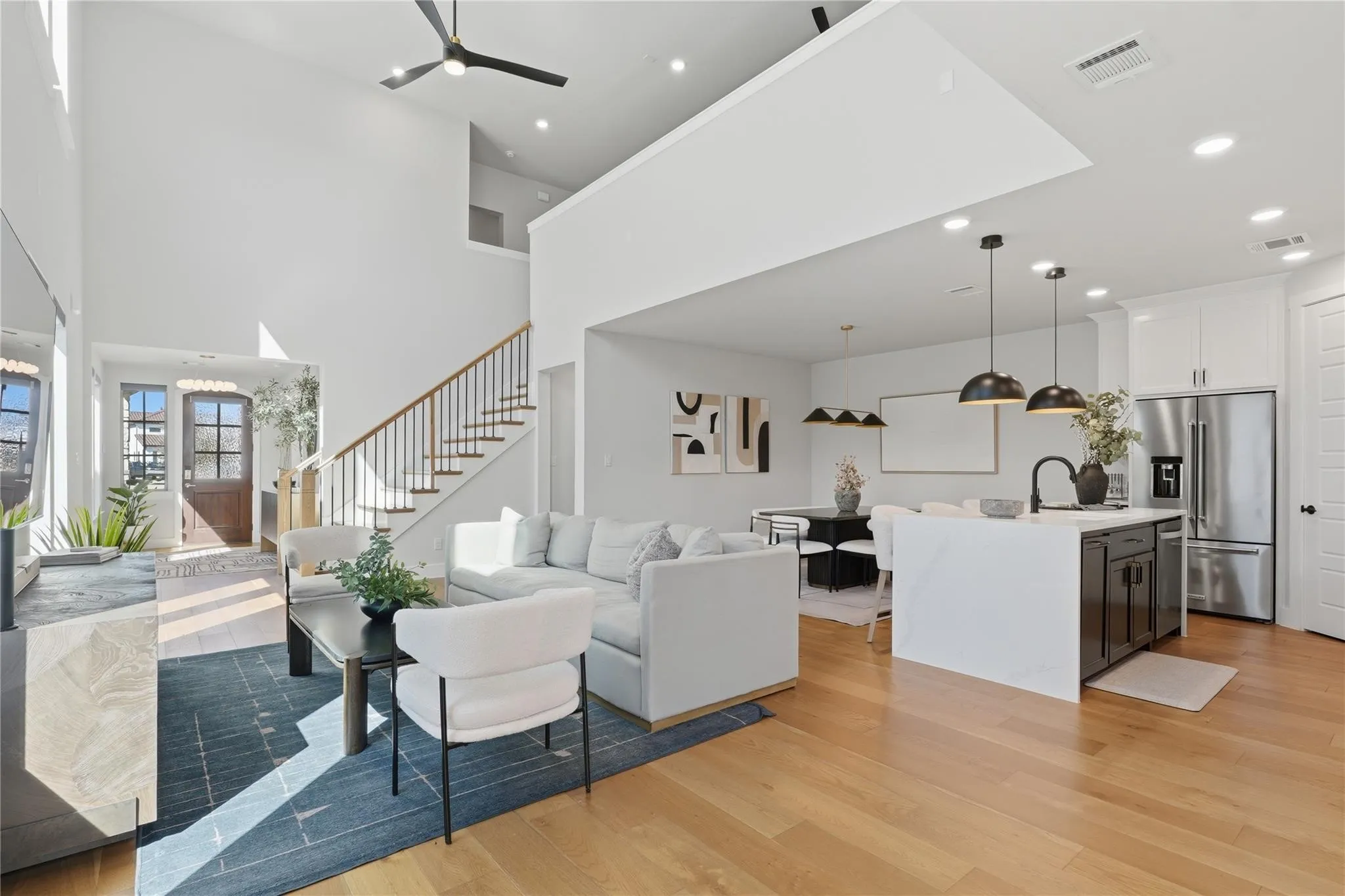 Open-concept living space featuring high ceilings, wood-finish flooring, a contemporary kitchen island with pendant lighting, and a prominent staircase with wood handrails
