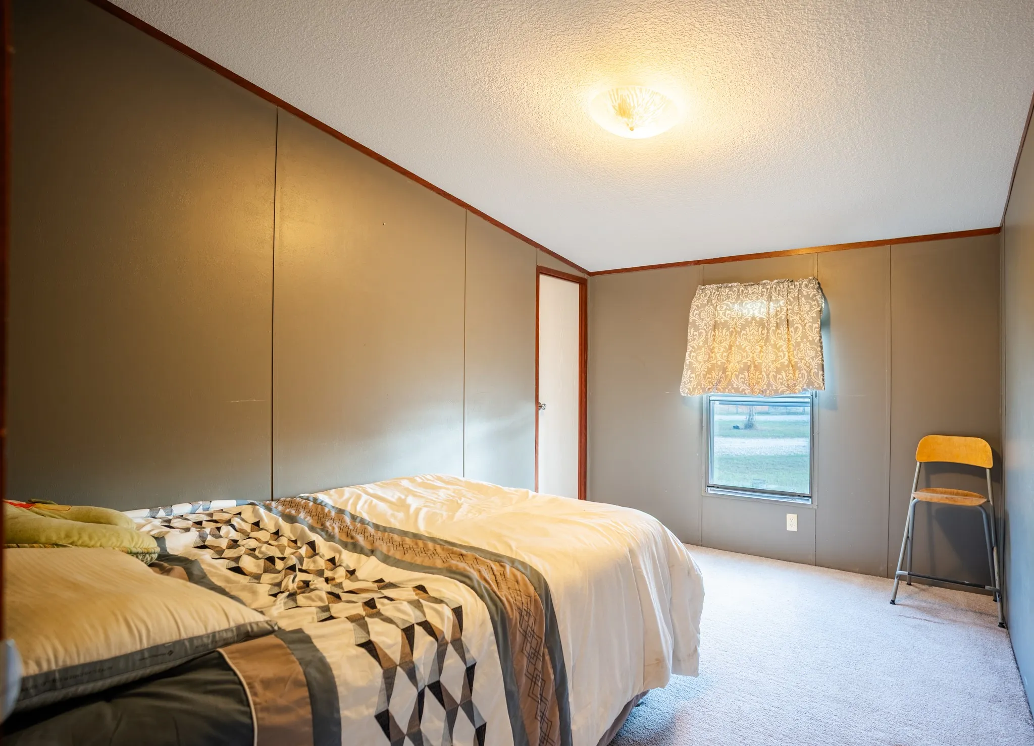 Wall paneling, light carpet flooring, ceiling-mounted light fixture, and a window with a valance