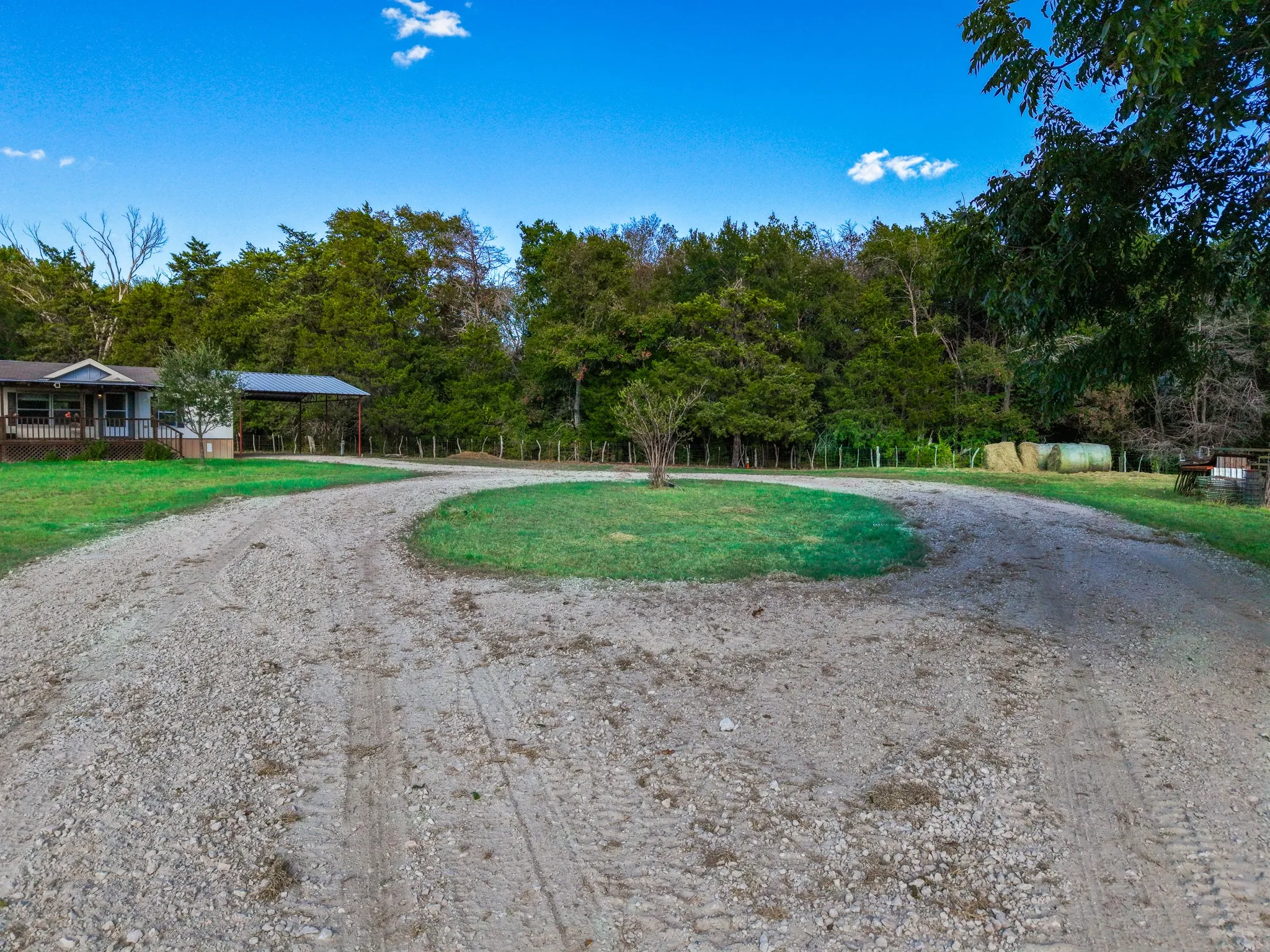 Gravel driveway with a central grassy island, mature trees, and a detached carport